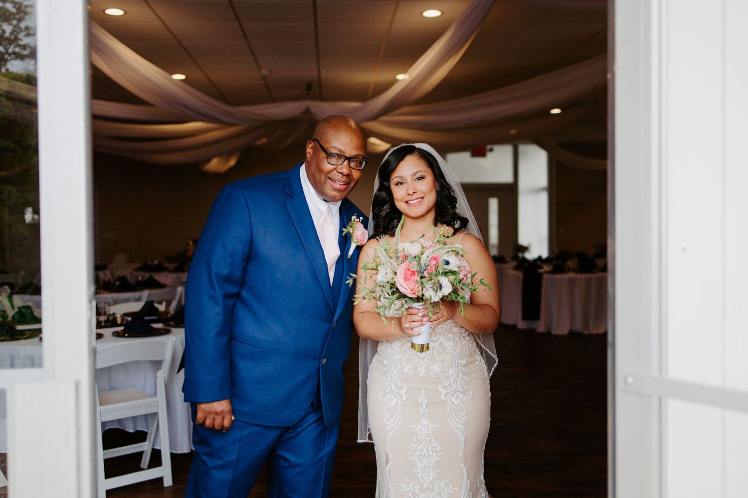 Bride in lace gown holds pink bouquet beside escort in blue suit at reception entrance.