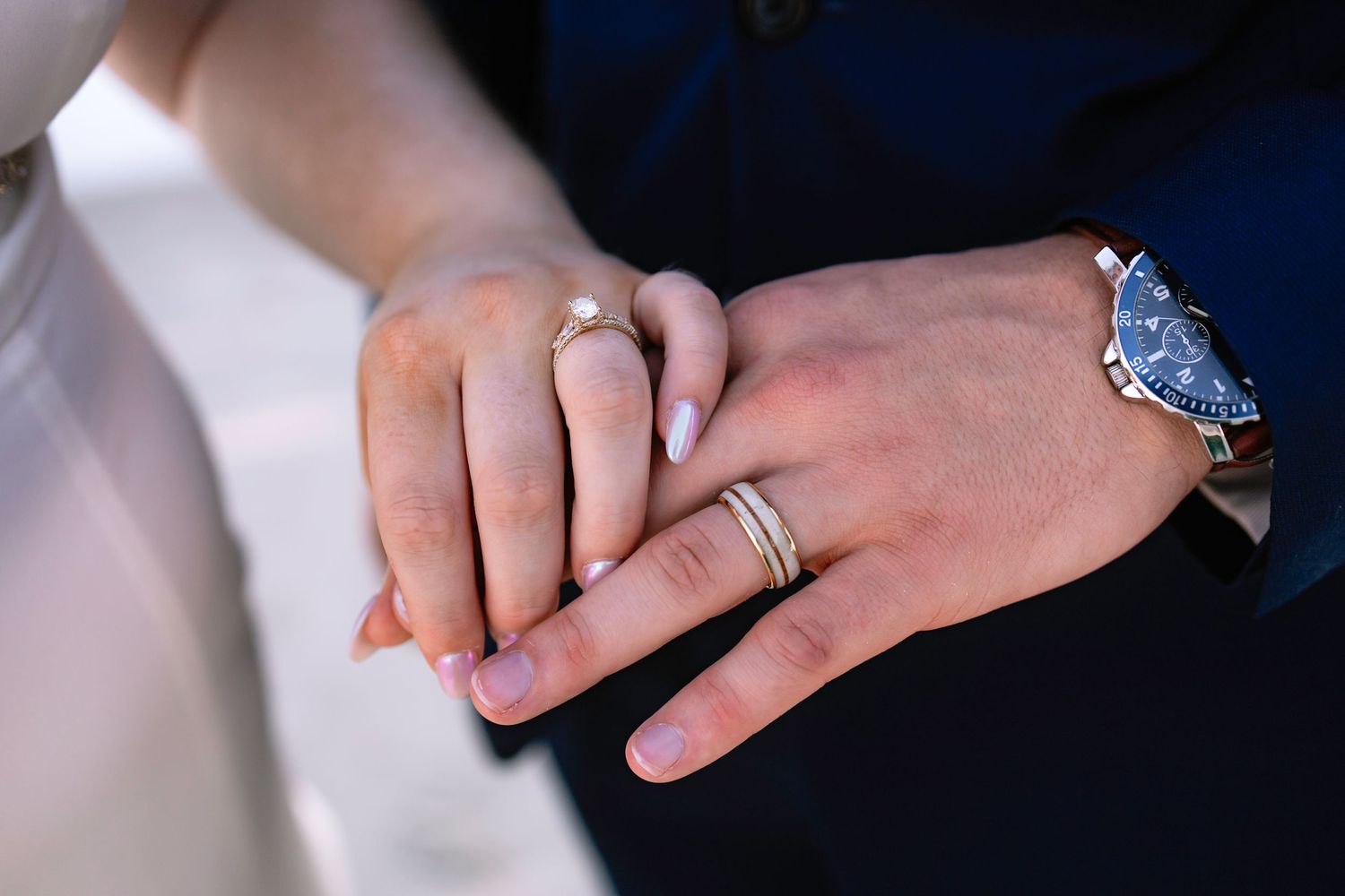 Close-up of bride and groom holding hands showing wedding rings and a blue watch near a white dress.