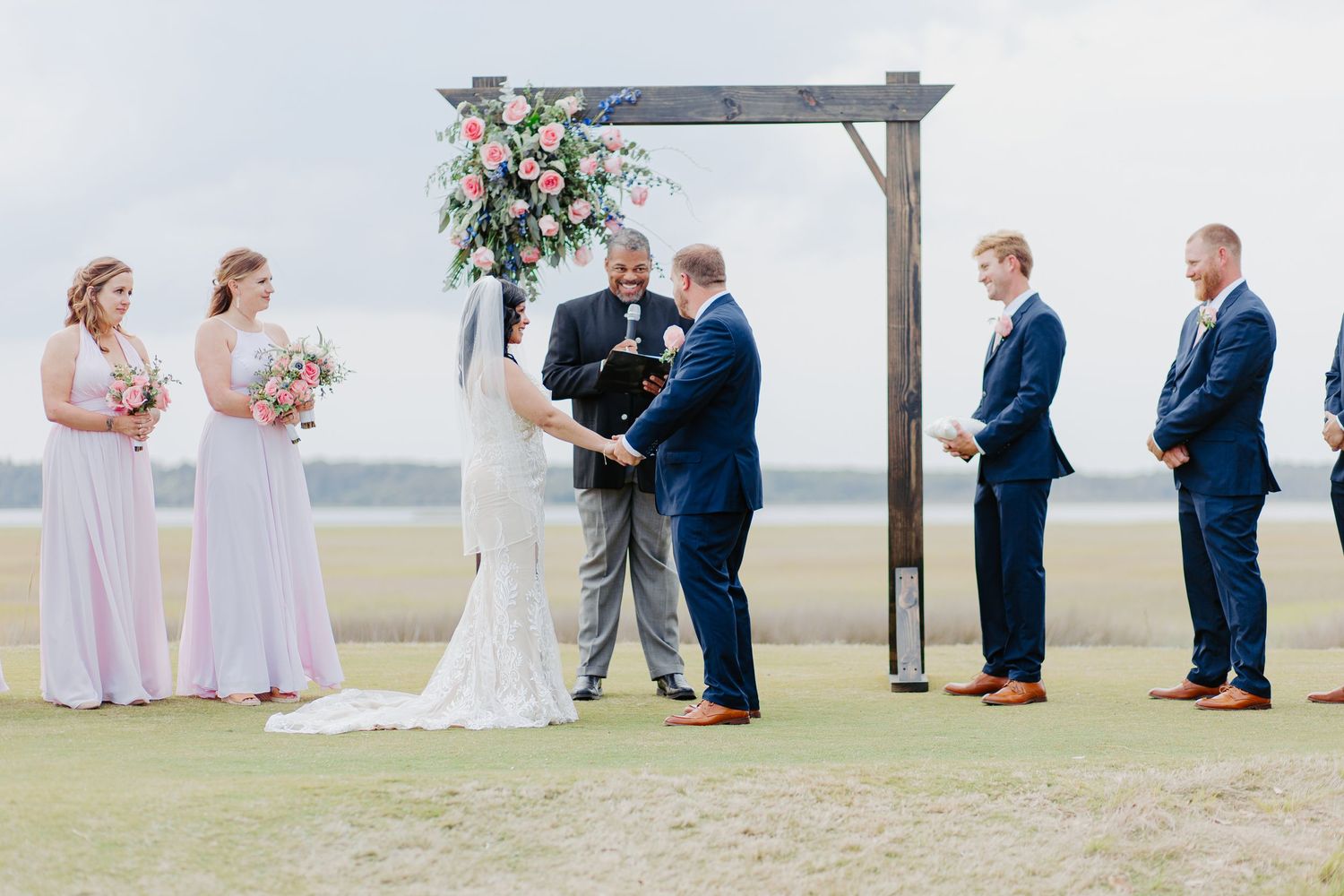 Couple exchanges vows at wooden arch with pink floral decor on scenic waterfront lawn.