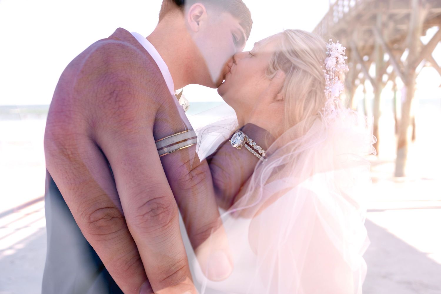 Bride and groom sharing a tender kiss near a beach pier with soft golden sunlight and flowing veil.
