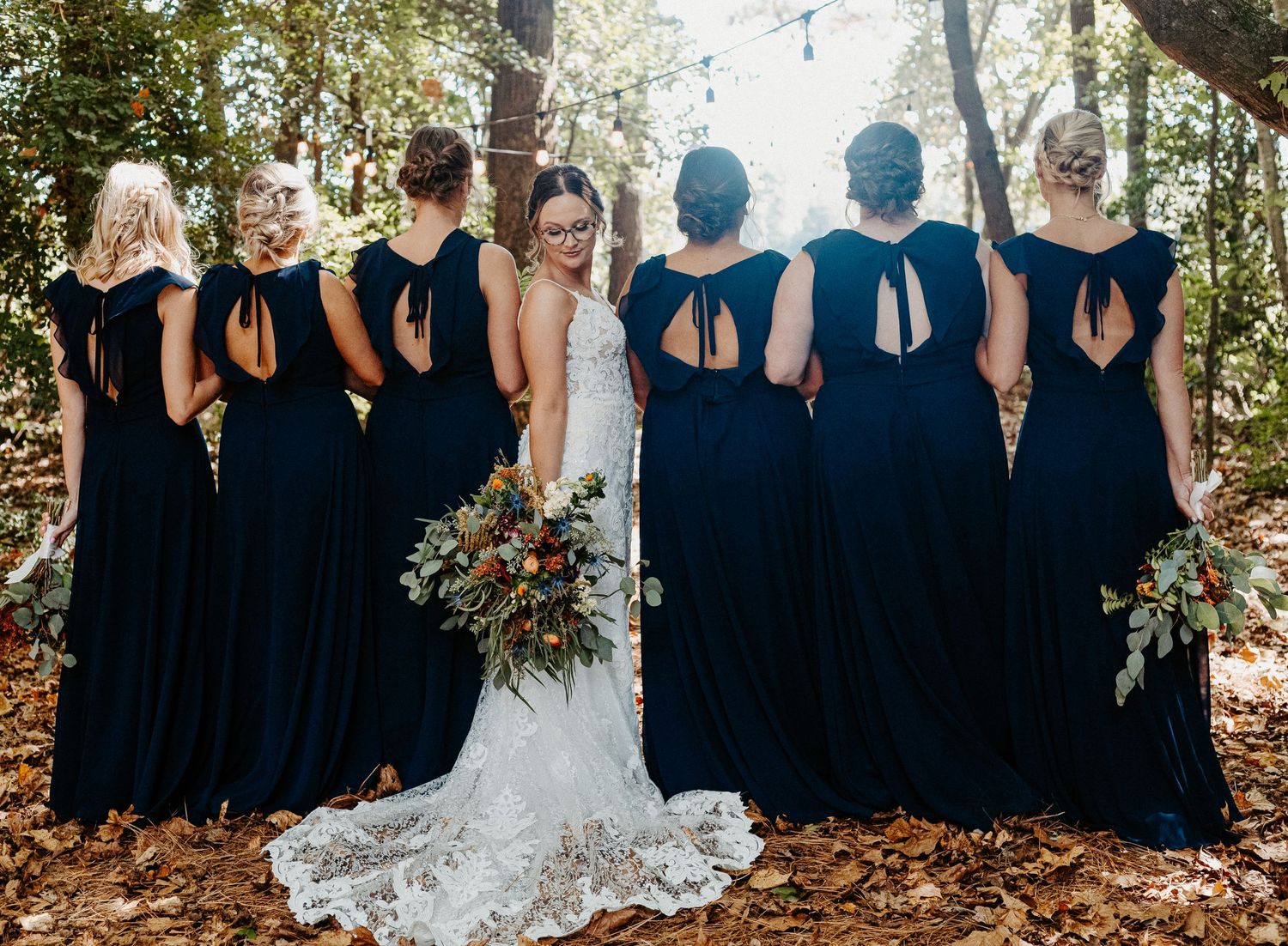 A bride in lace gown stands with bridesmaids in navy dresses holding bouquets in an autumn forest.