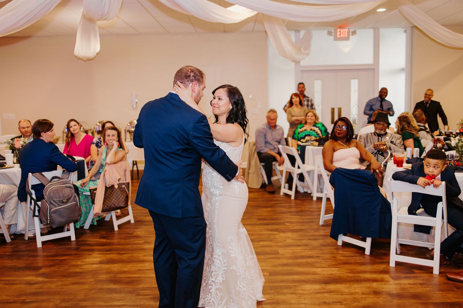 Bride and groom share first dance at wedding reception while guests watch from white chairs.