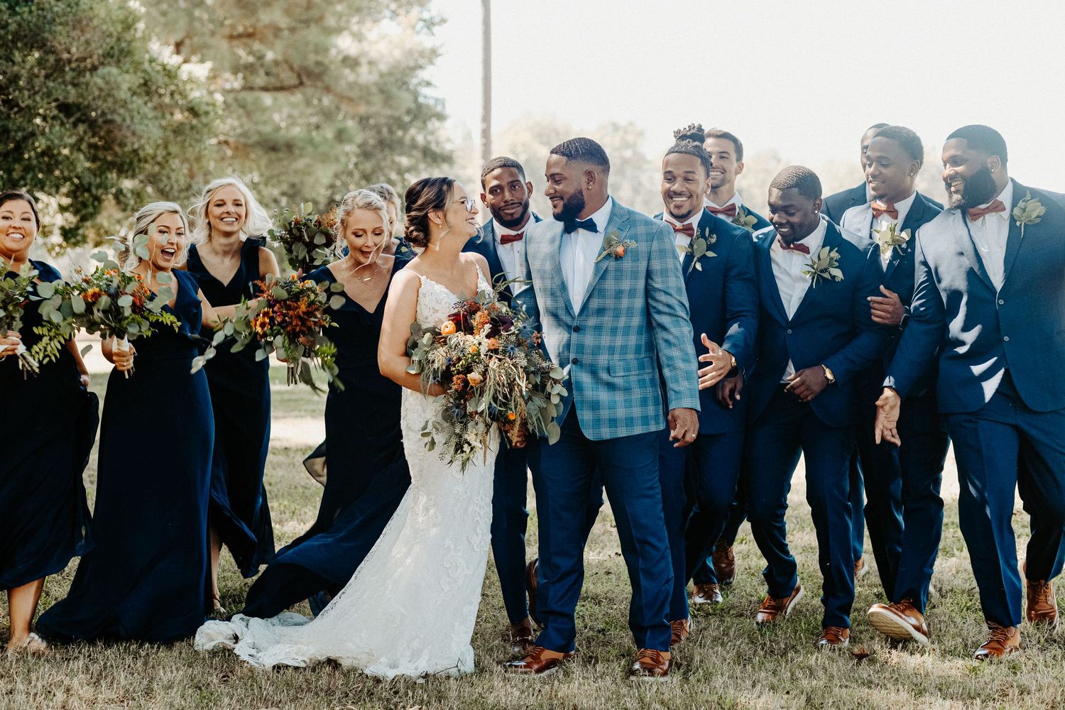 Wedding party laughs joyfully outdoors in navy suits and dresses with fall bouquets.