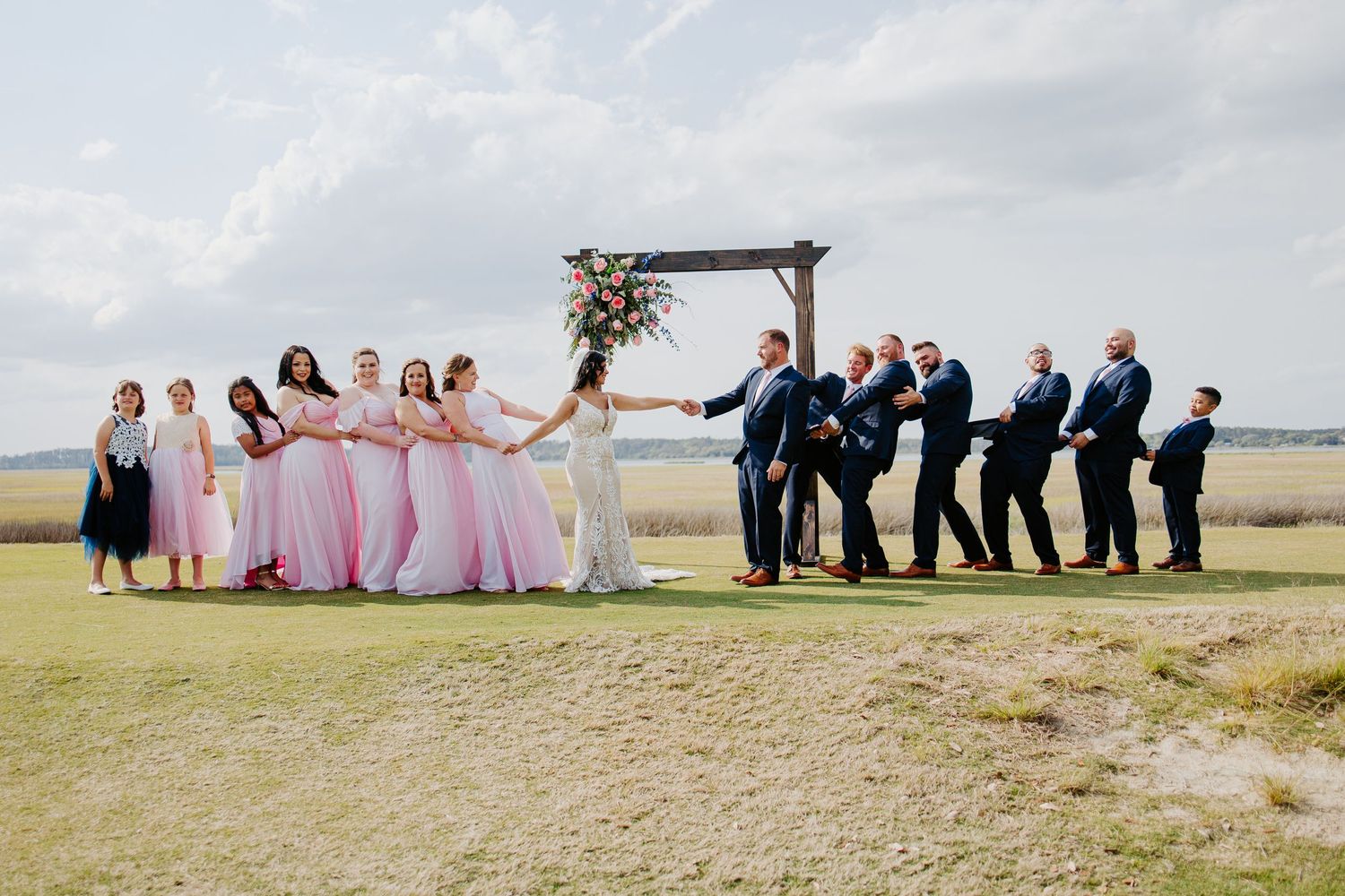 Wedding party lines up at outdoor wooden arch on open field in pink and navy attire.