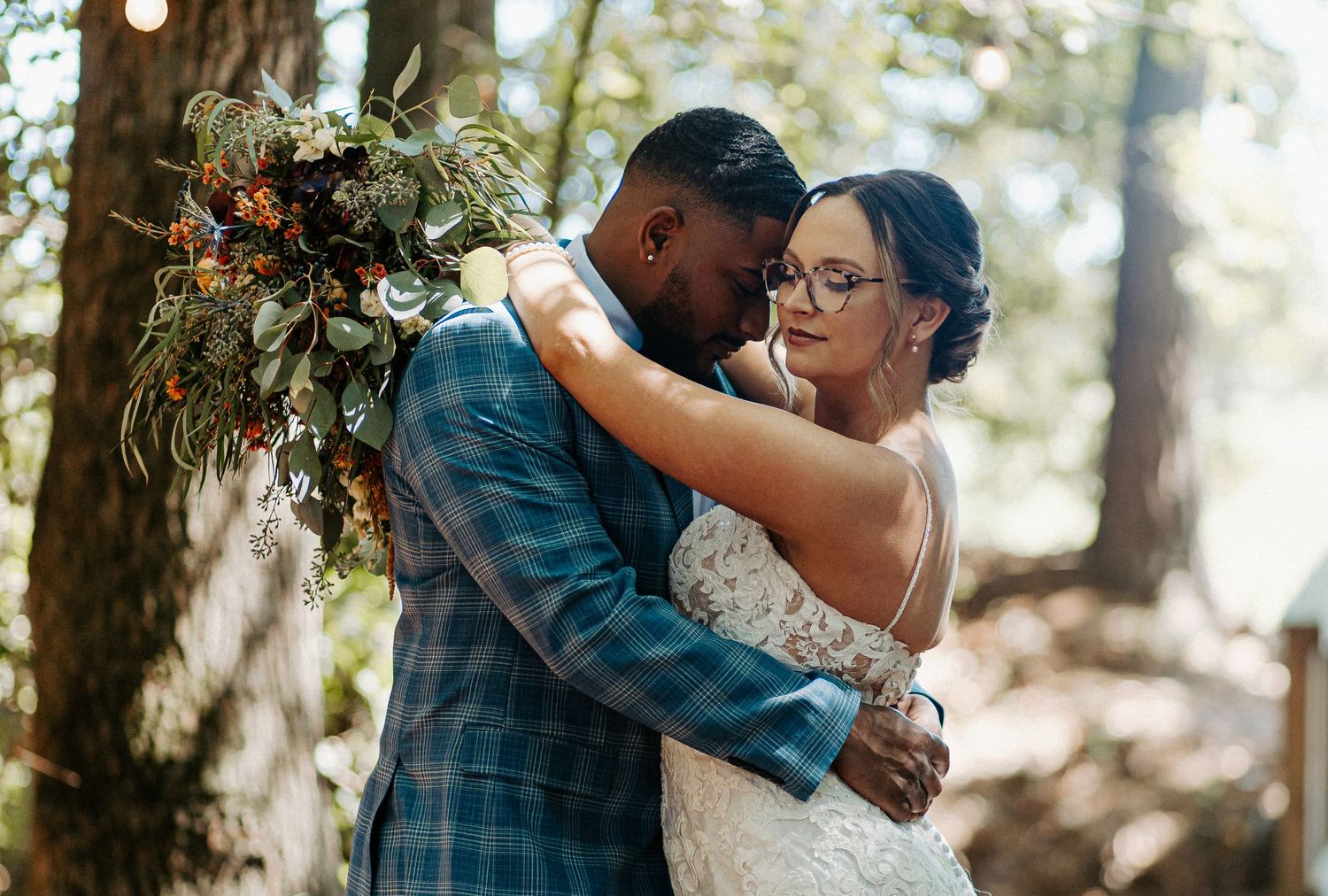 Bride and groom share intimate embrace in forest setting with lush bridal bouquet.