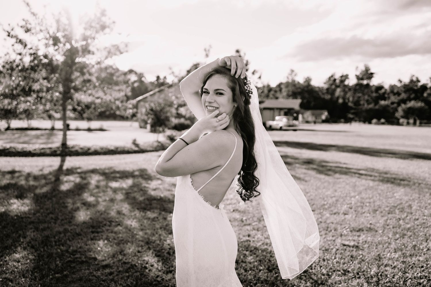 Black and white photo of a smiling bride in a backless wedding gown with veil flowing in the wind outdoors.