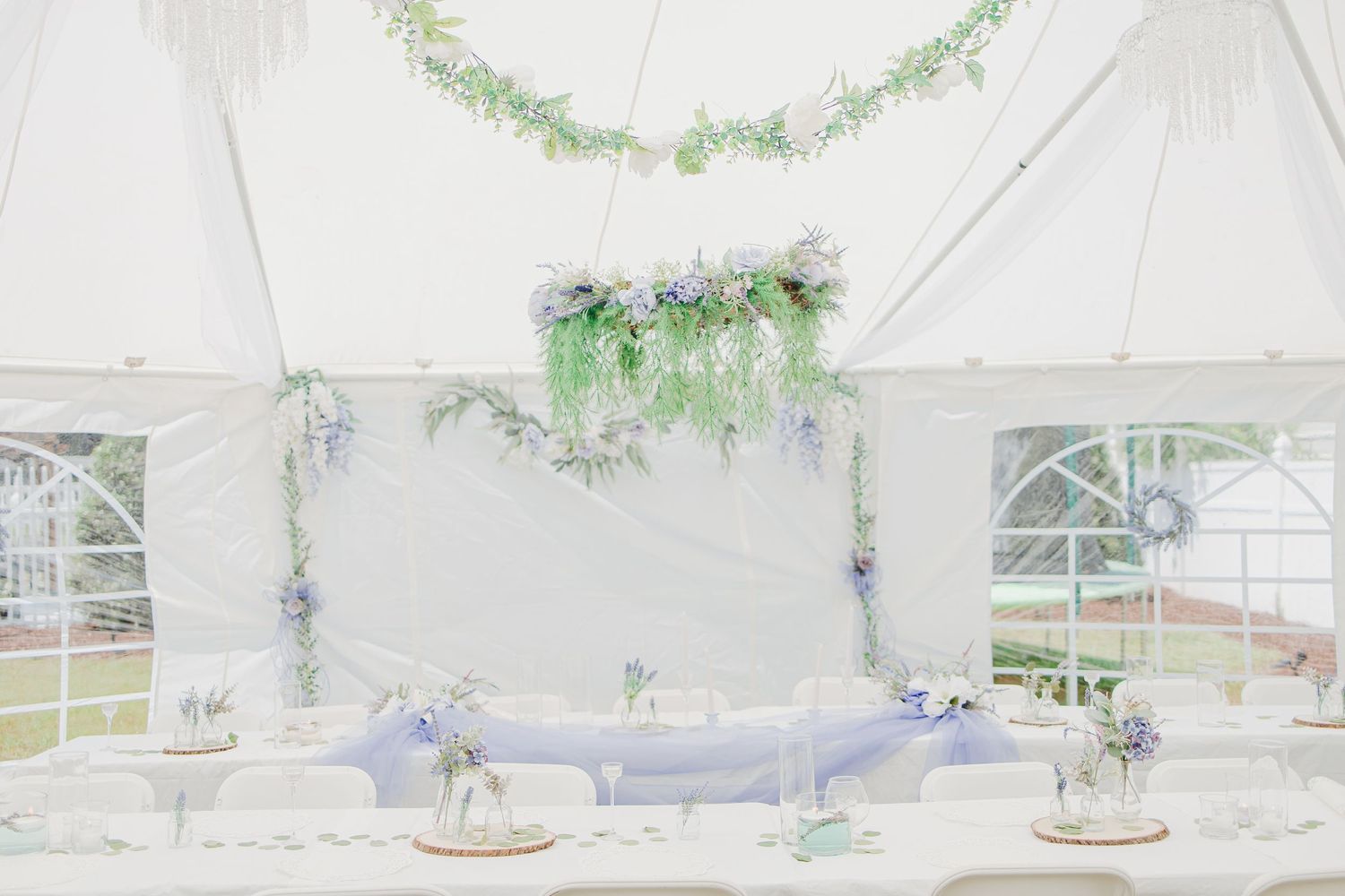 Elegant wedding tent reception with floral chandelier, lavender draping, and white table settings in soft pastel tones.
