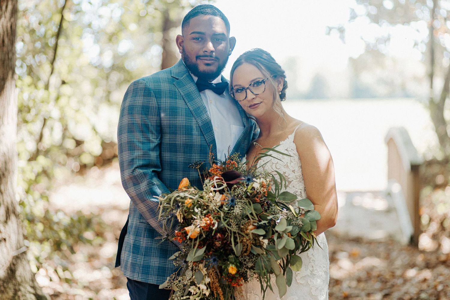 A smiling bride and groom pose together outdoors, with the groom in a plaid suit and bride holding a lush bouquet.