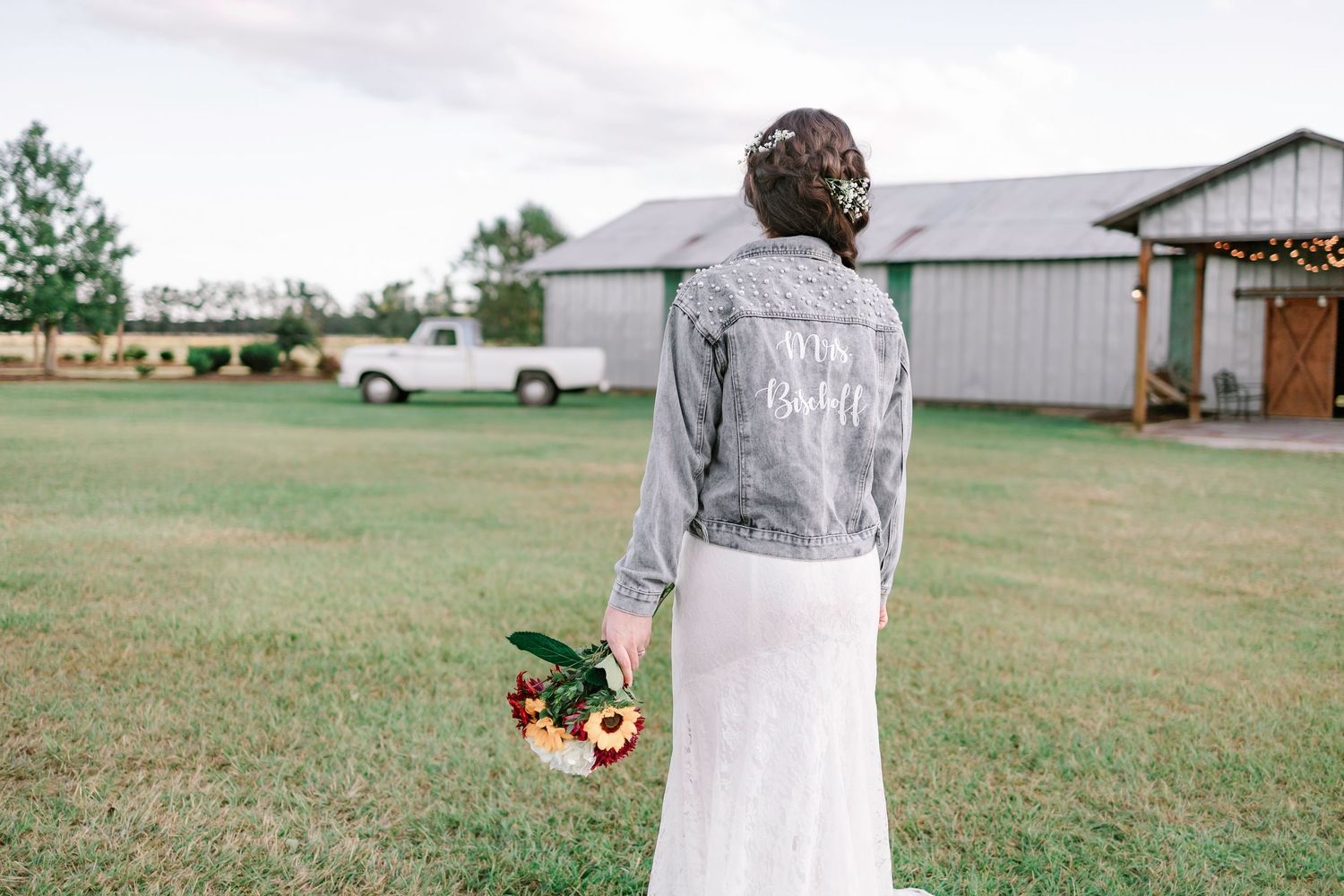 Bride in lace dress and custom denim jacket holding colorful bouquet at rustic farm wedding venue.