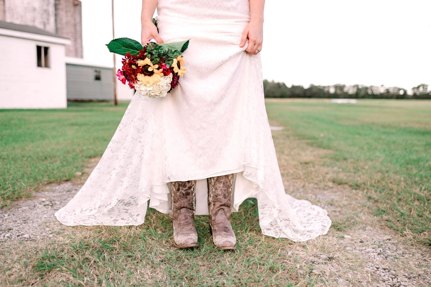 Bride in lace wedding dress and cowboy boots holding red and white bouquet on green lawn.