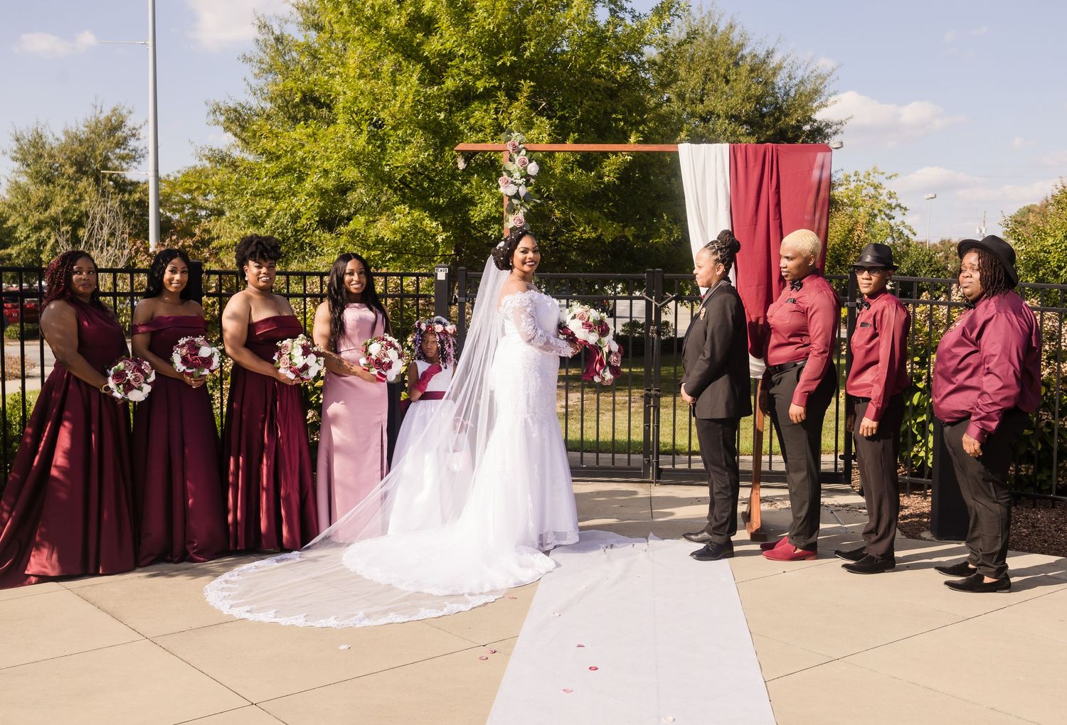 A bride and wedding party pose outdoors near a floral arch in burgundy and pink attire.