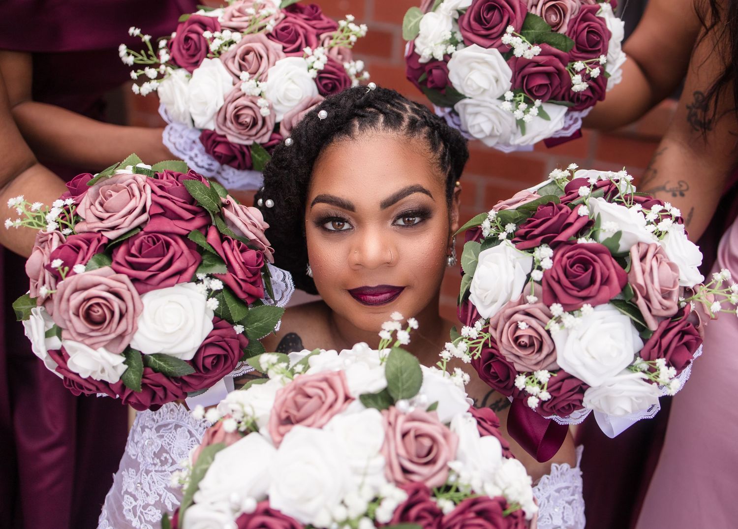 A bride surrounded by bridesmaids holding burgundy, pink, and white rose bouquets.