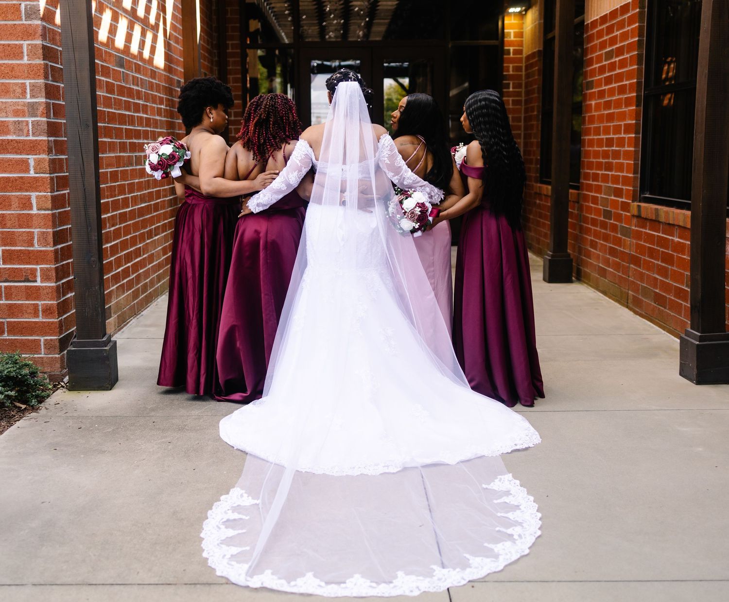 A bride in a white gown with a long lace veil stands with burgundy-dressed bridesmaids.