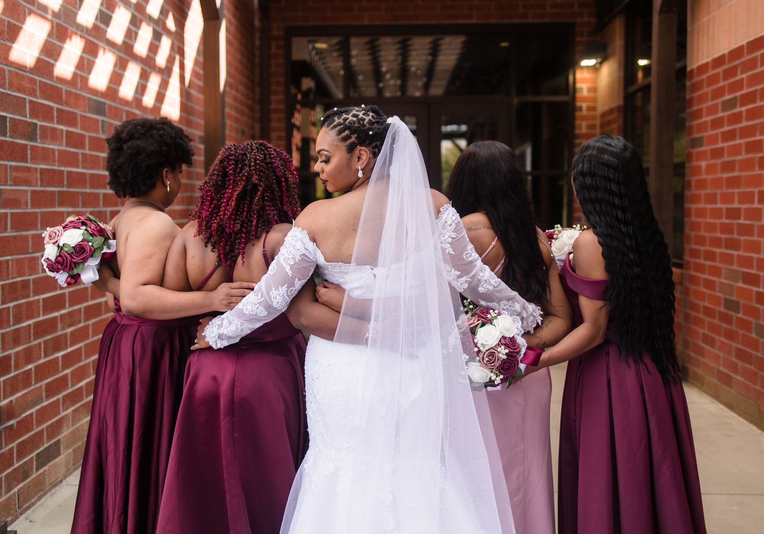 A bride in a white lace wedding gown embraces her bridesmaids in burgundy satin dresses.