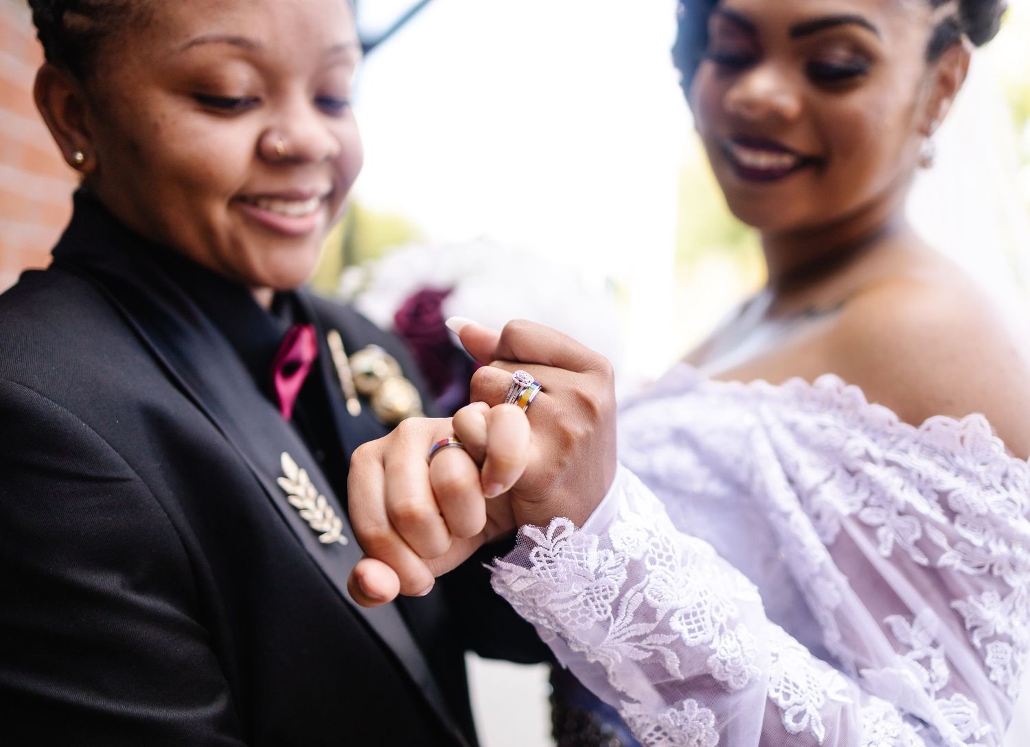 Two brides exchange rings during their wedding ceremony, one in a black suit and one in a white lace gown.