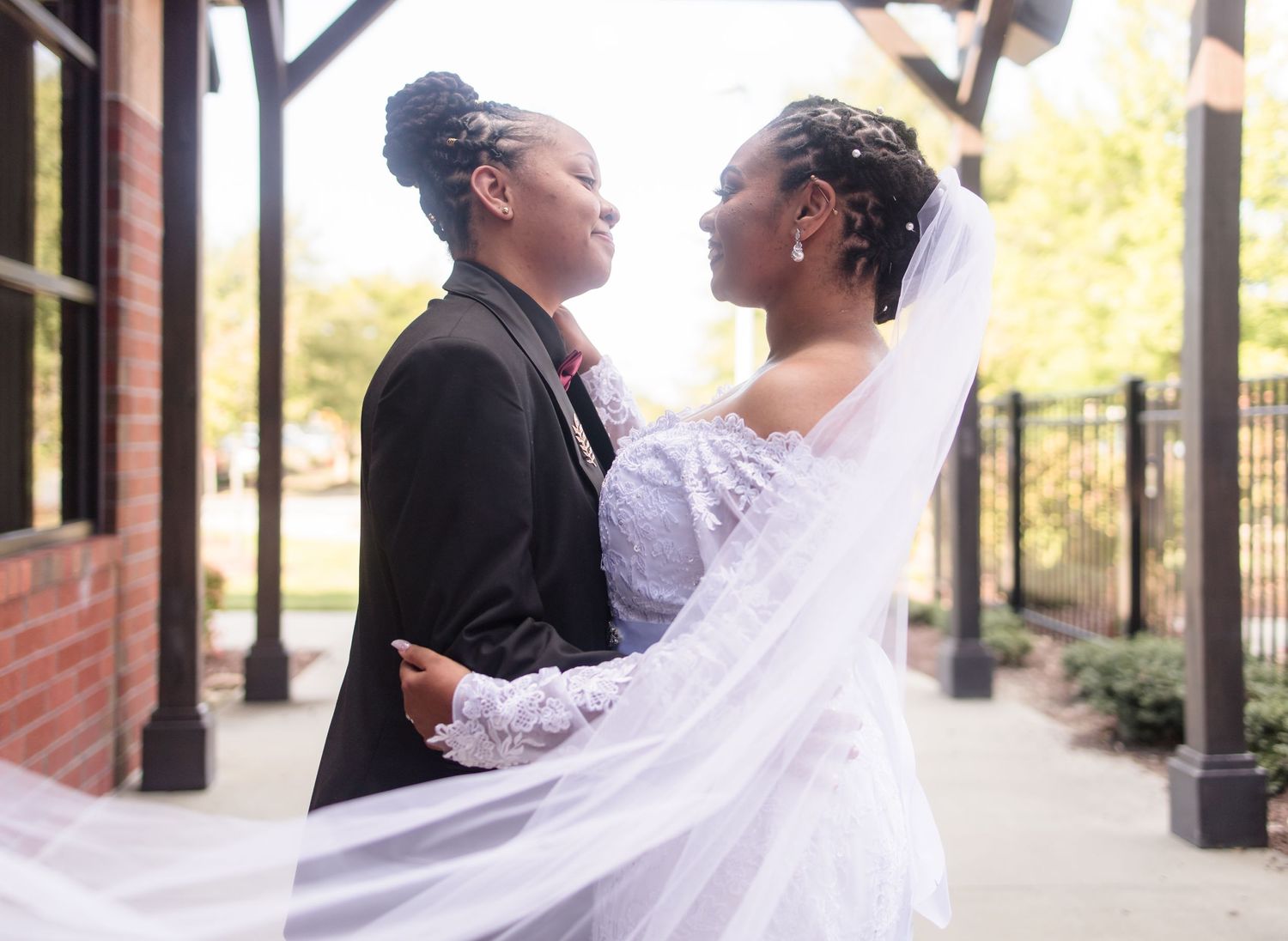 Two brides share an intimate moment outdoors, one in a white wedding gown with veil and one in a black suit.
