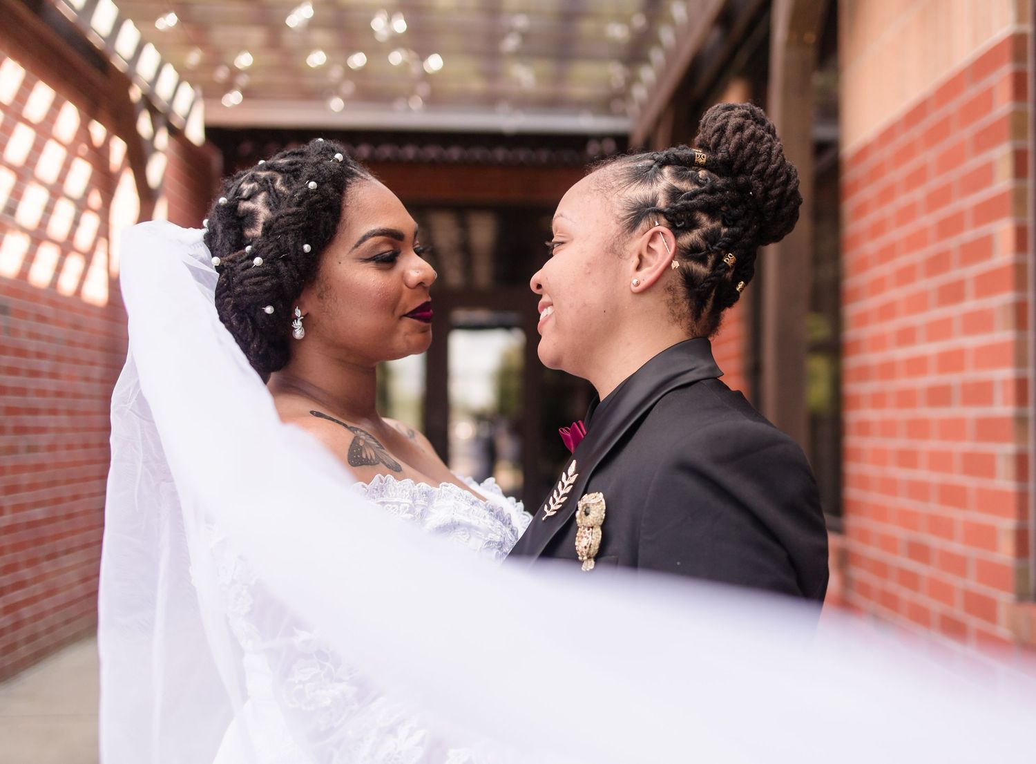 Two brides gaze lovingly at each other, one in a white lace wedding dress with veil and one in a black suit.