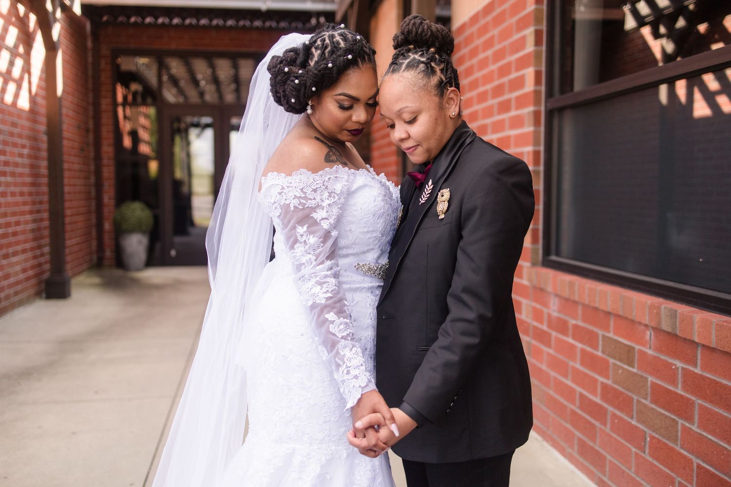Two brides hold hands tenderly near a brick wall, one in a white off-shoulder gown and one in a black suit.