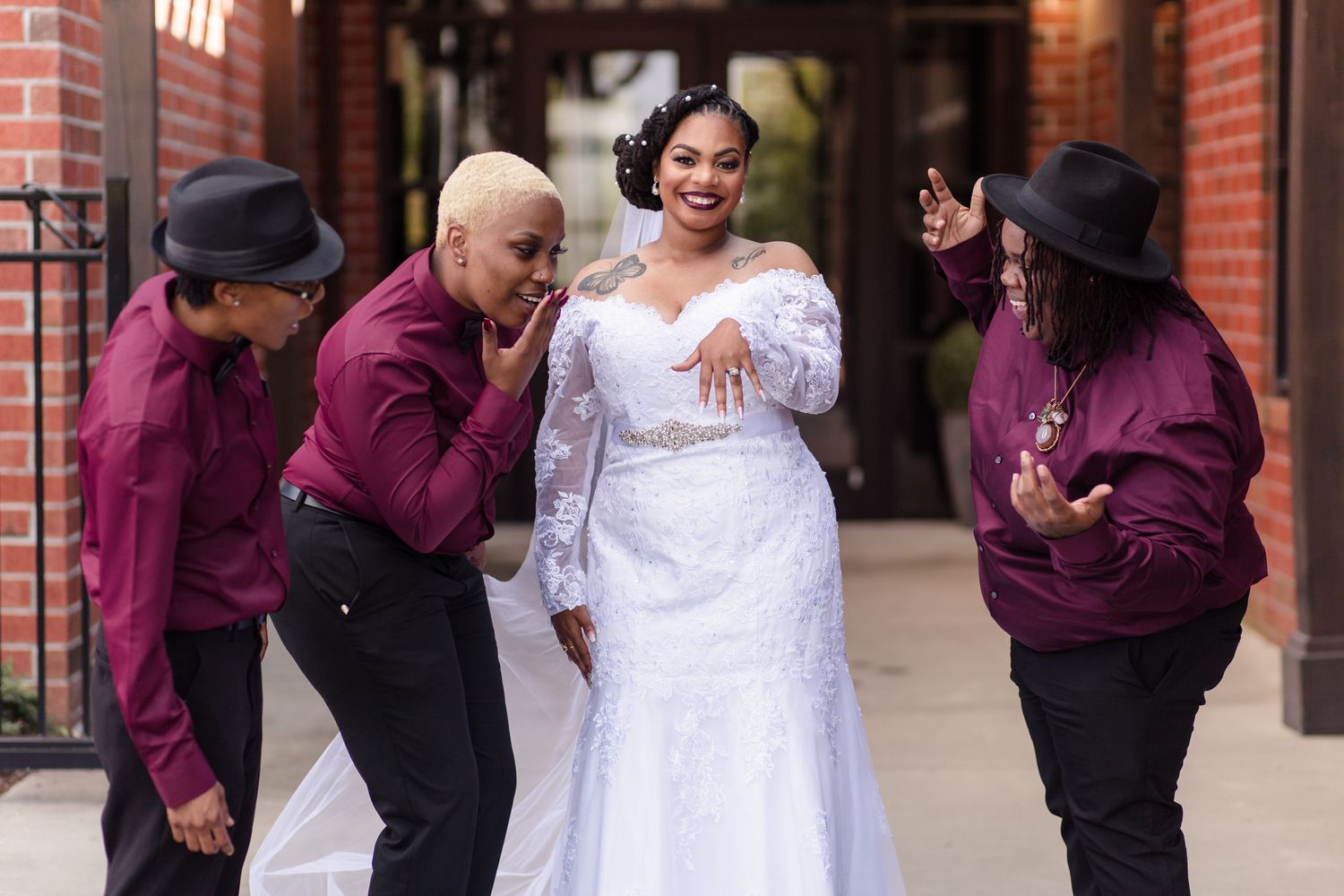 A bride in a white lace gown laughs joyfully with three bridesmaids in burgundy outfits outdoors.