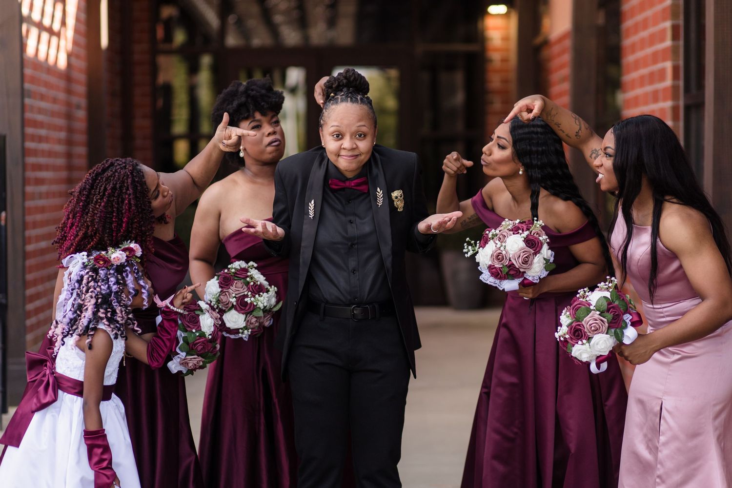 A person in a black suit walks joyfully surrounded by bridesmaids in burgundy dresses holding bouquets.