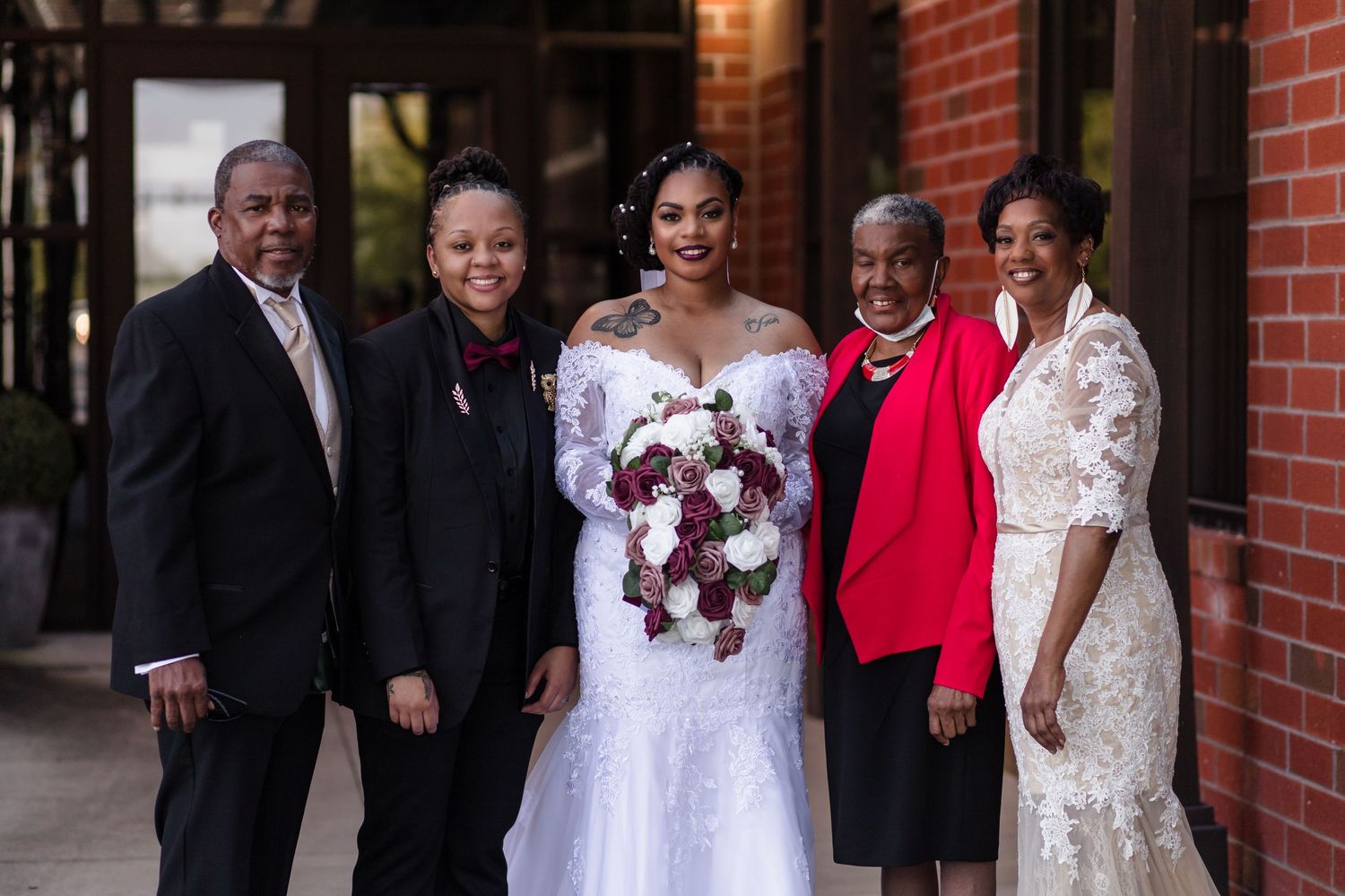 A bride in a white gown poses with four family members in front of a brick building.