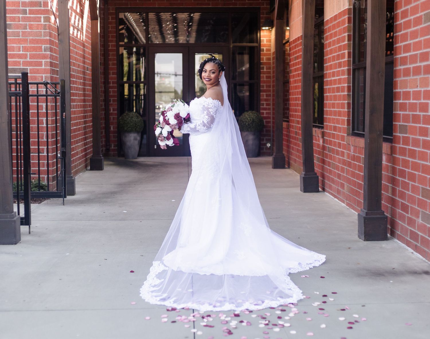 Bride in white lace gown with long veil holds bouquet outside brick building.