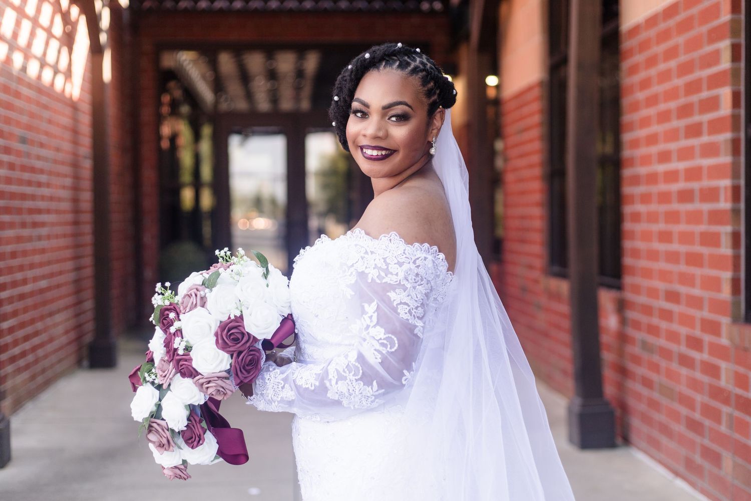 Smiling bride in off-shoulder lace wedding dress holds purple and white bouquet.