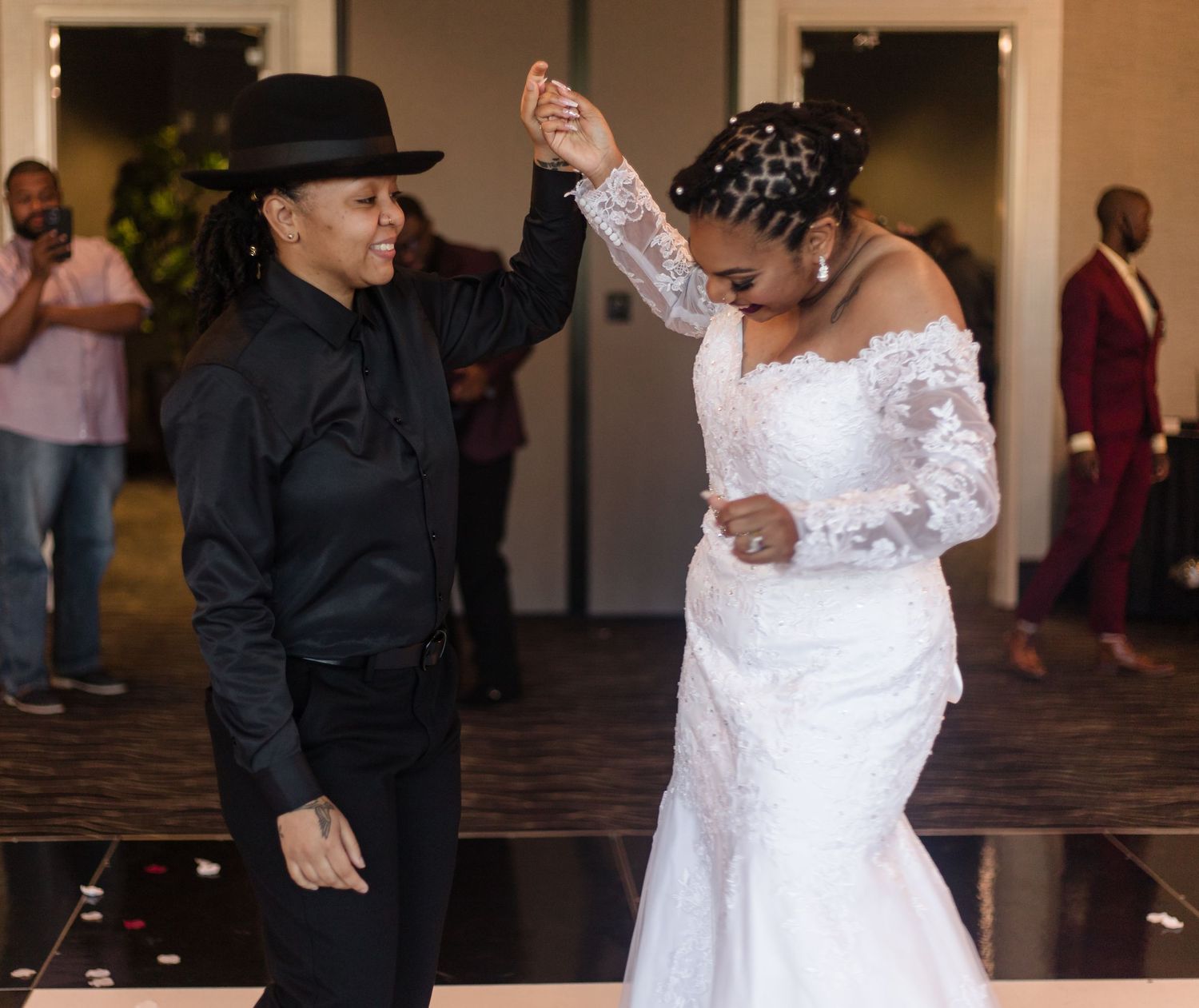 Two brides dance joyfully, one in white gown and one in black suit with hat.