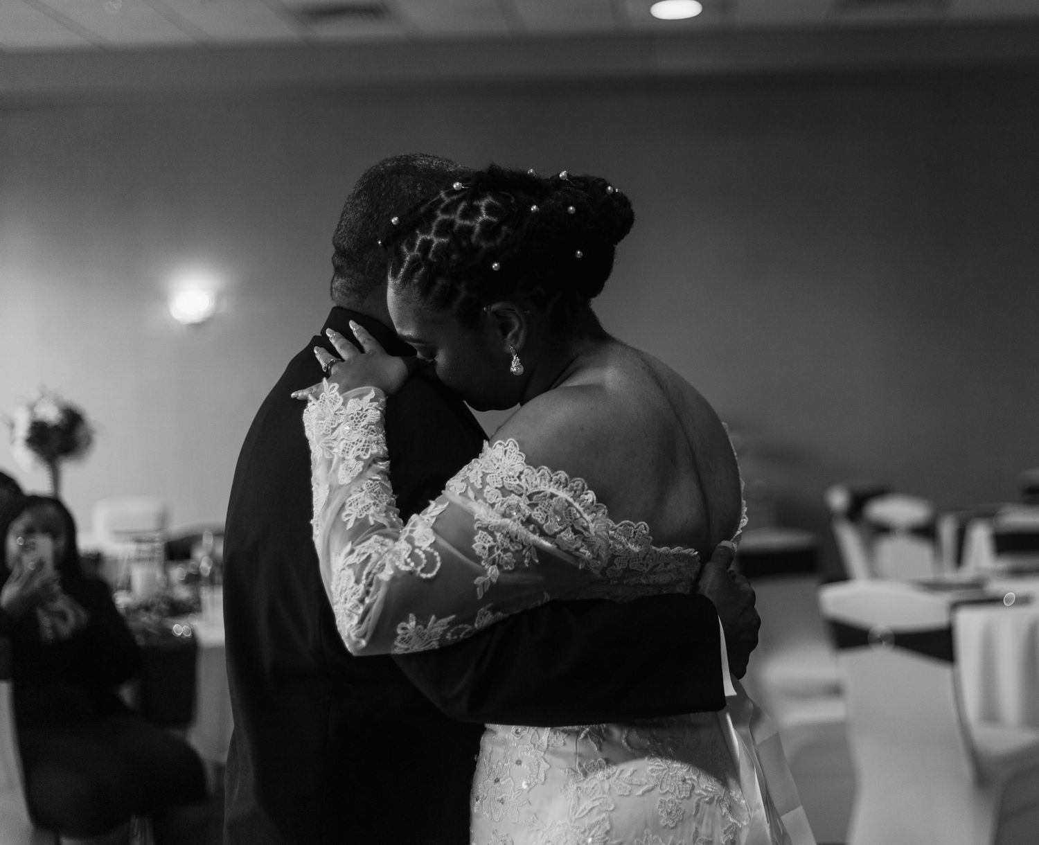 Bride in lace gown shares tender embrace during slow dance at wedding reception.