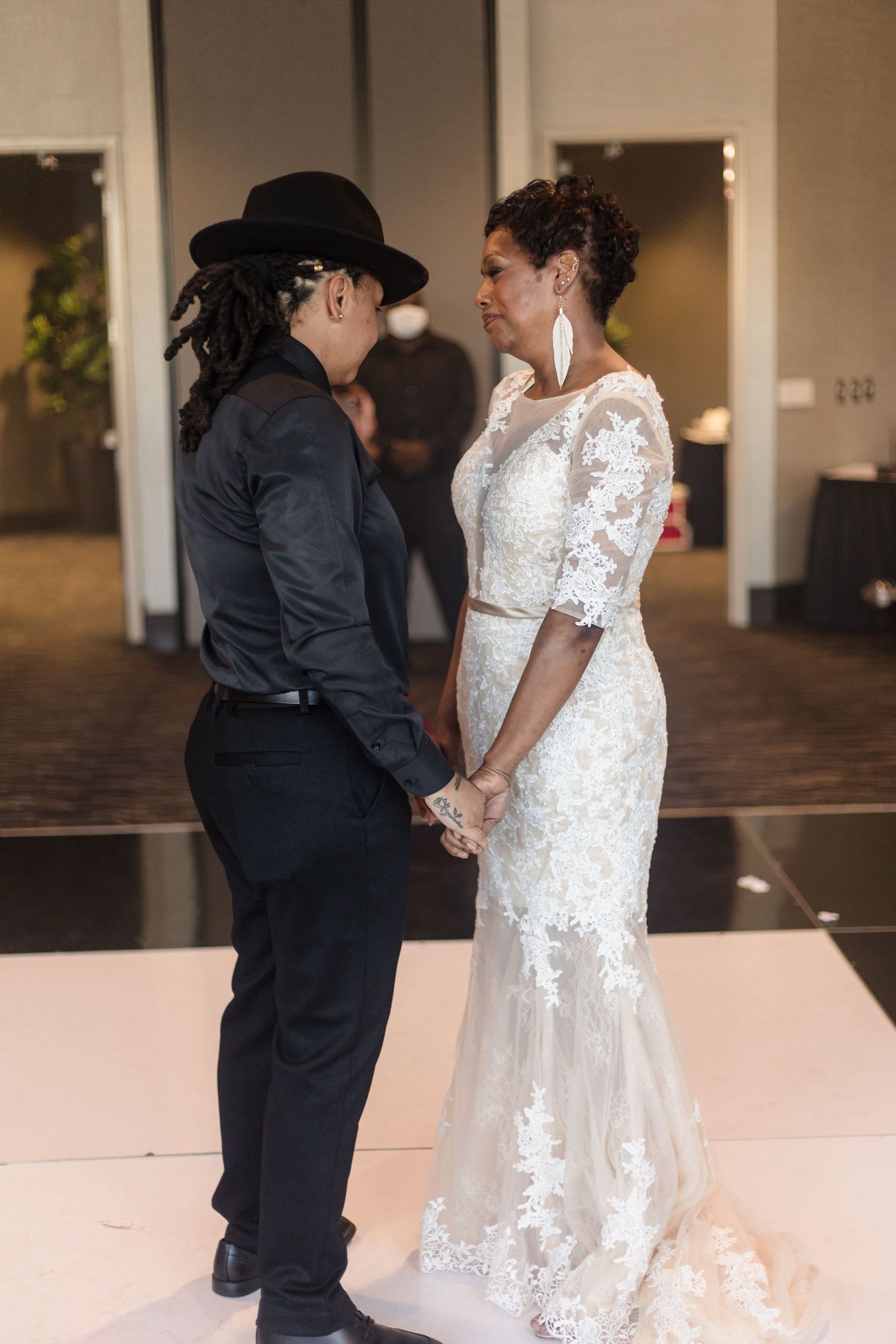 Two women hold hands on a white dance floor, one wearing a lace wedding gown at their wedding.