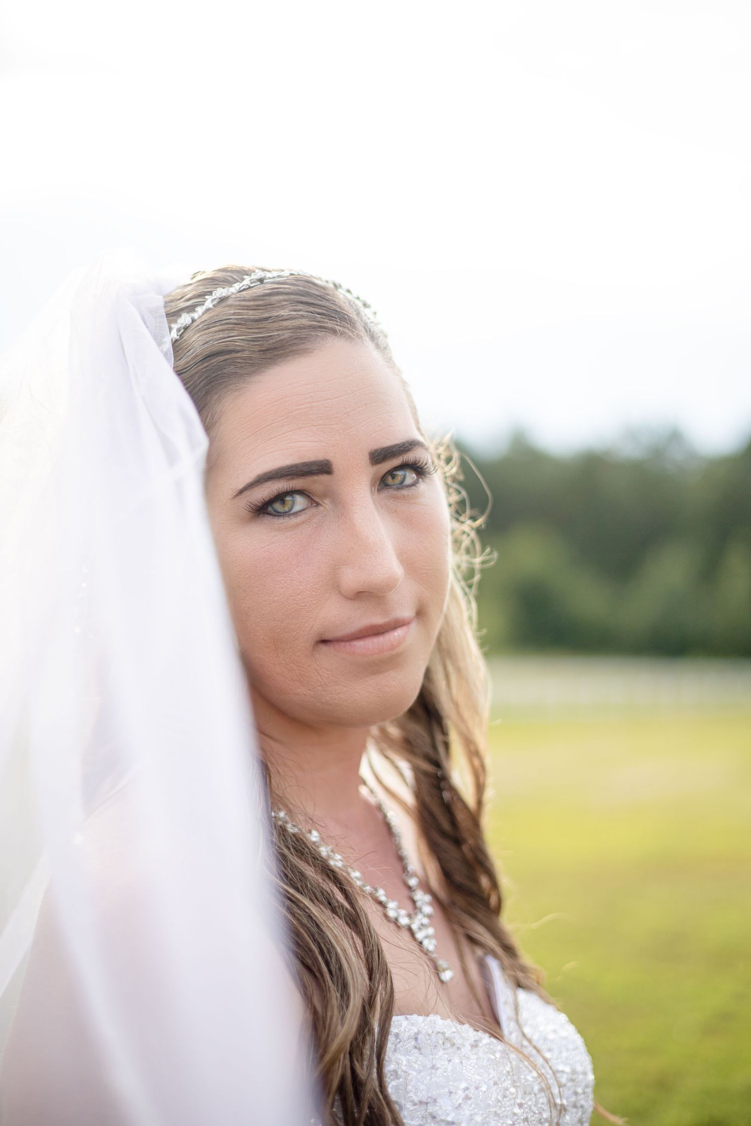 Beautiful bride in wedding dress and veil with crystal necklace posing in outdoor field.