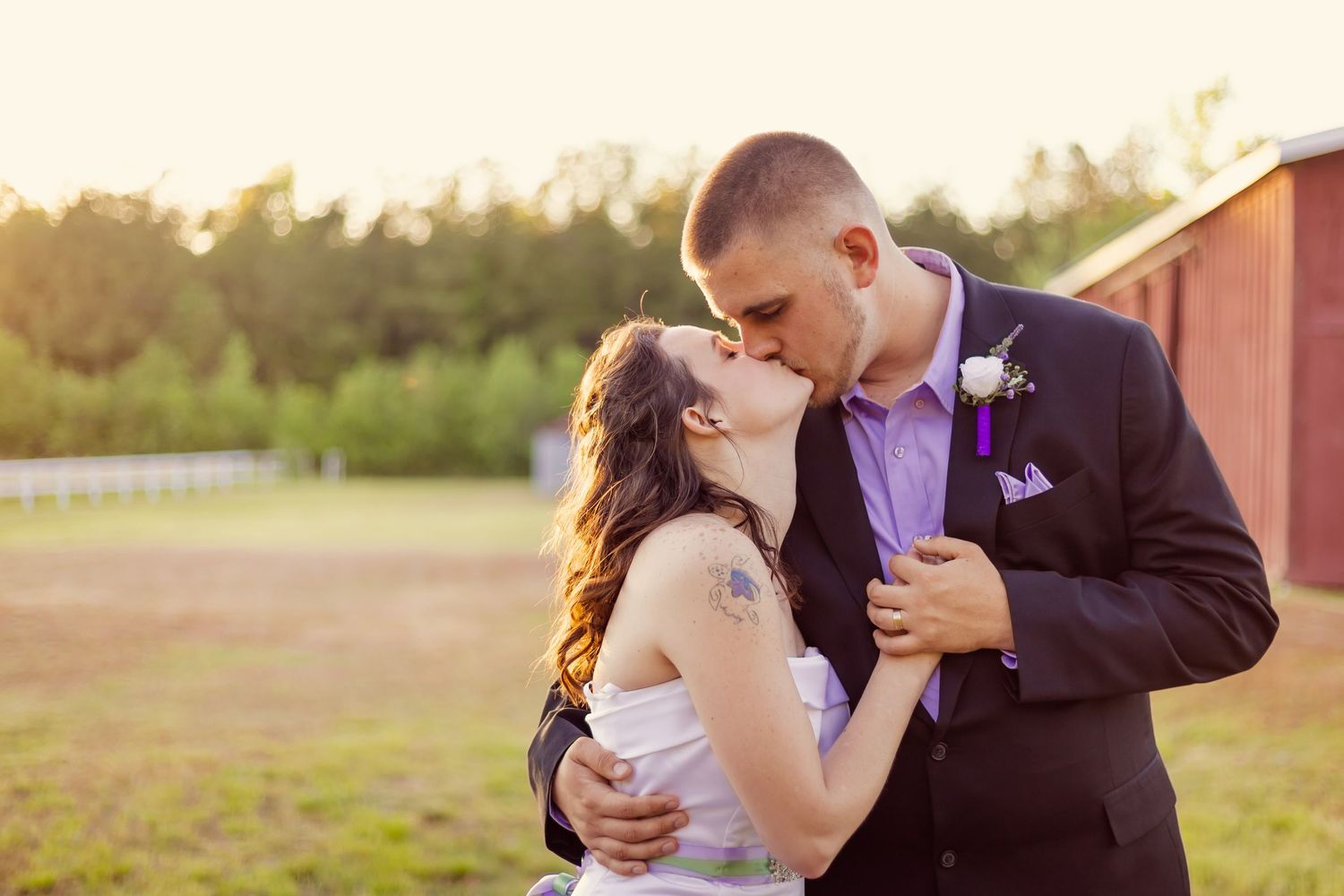 Bride and groom share a romantic kiss outdoors at golden hour near a red barn at their wedding.