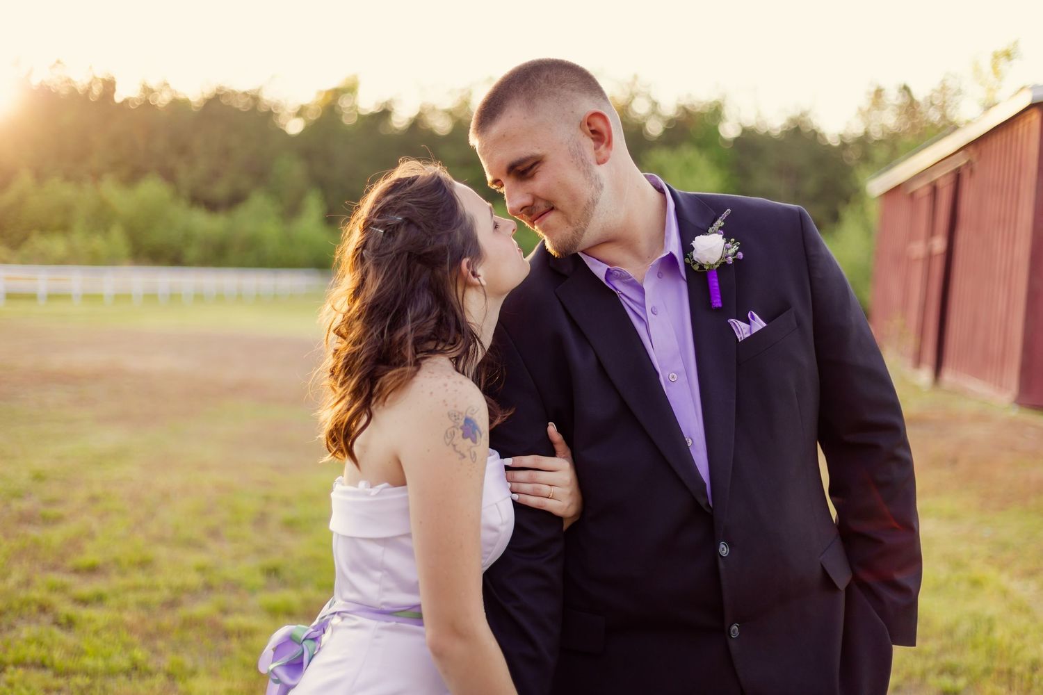Bride and groom sharing tender moment at golden hour near white fence with red barn backdrop.
