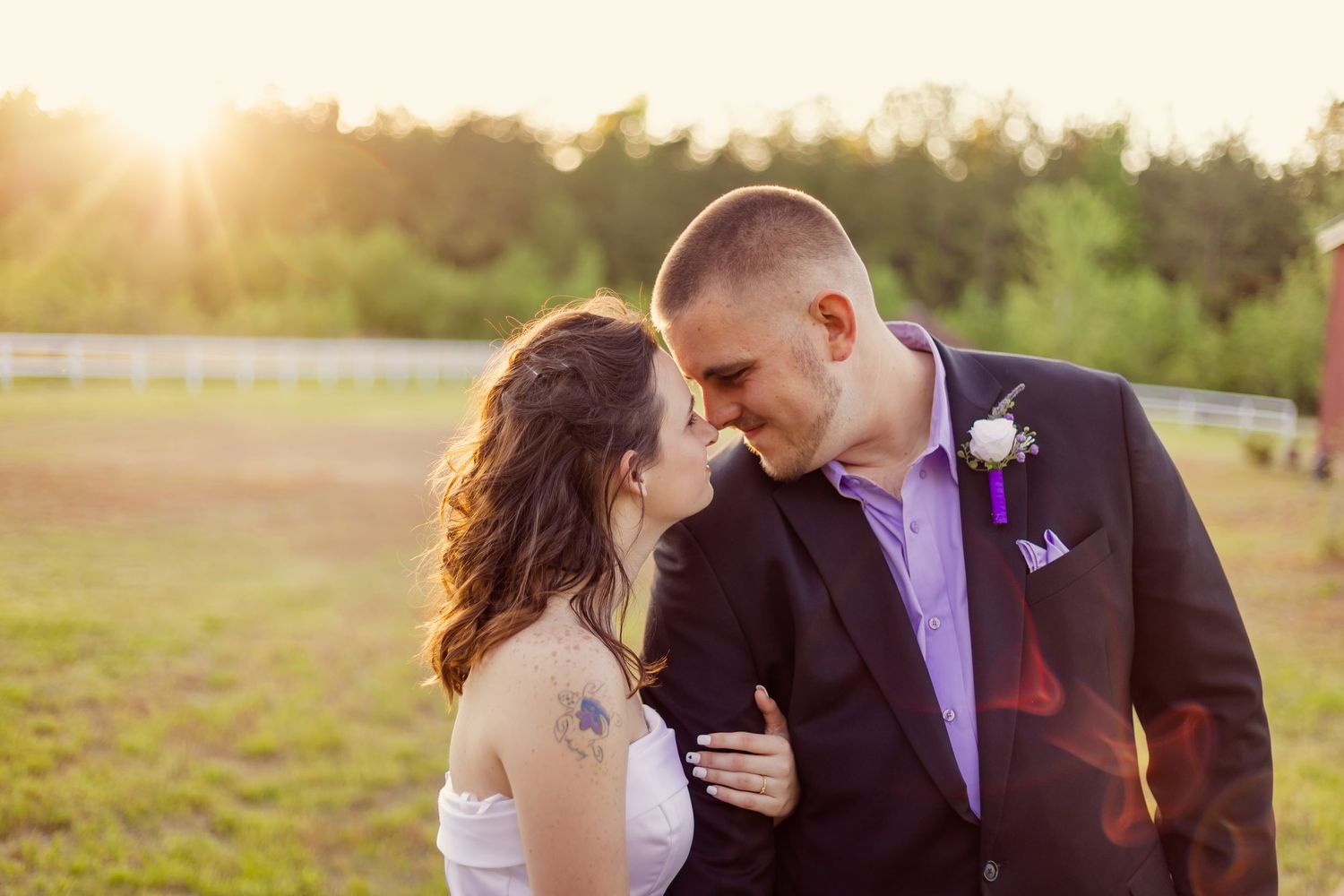 Wedding couple gazing at each other romantically at sunset near white fence and red barn outdoors.