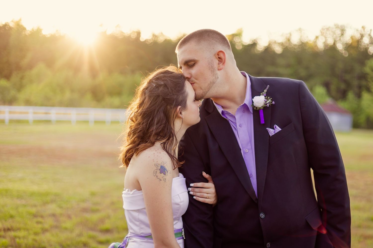 Wedding couple kissing in a sunlit field at sunset, groom in navy suit with purple boutonniere.