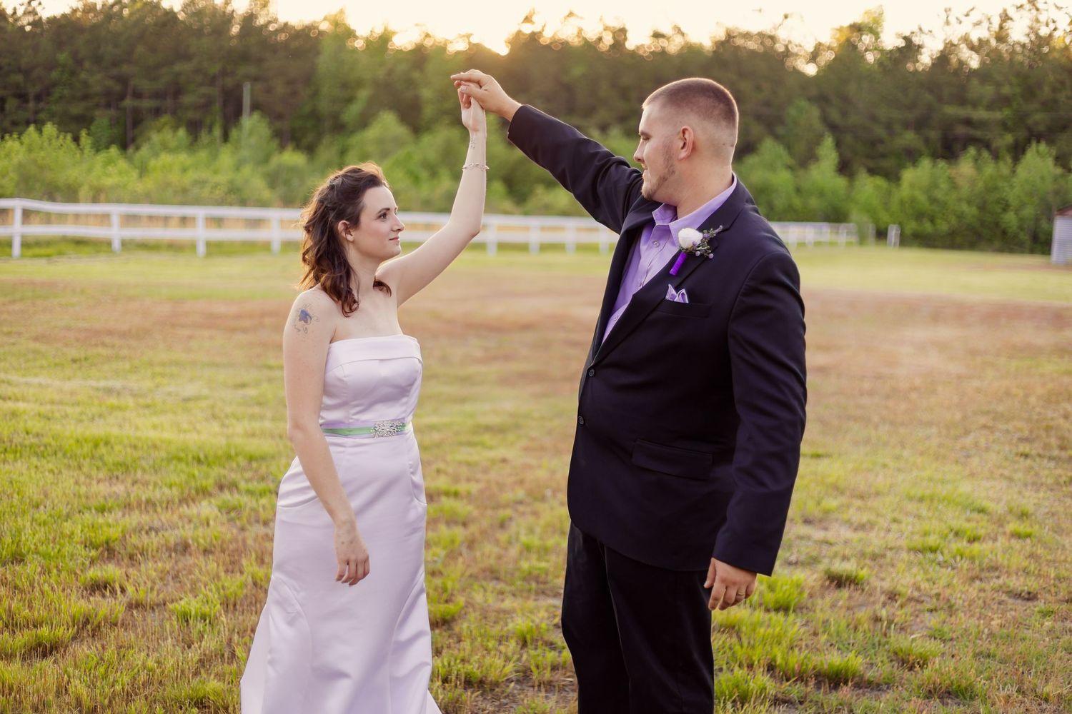 Groom twirls bride in white wedding gown on green field during golden hour outdoor photo session.