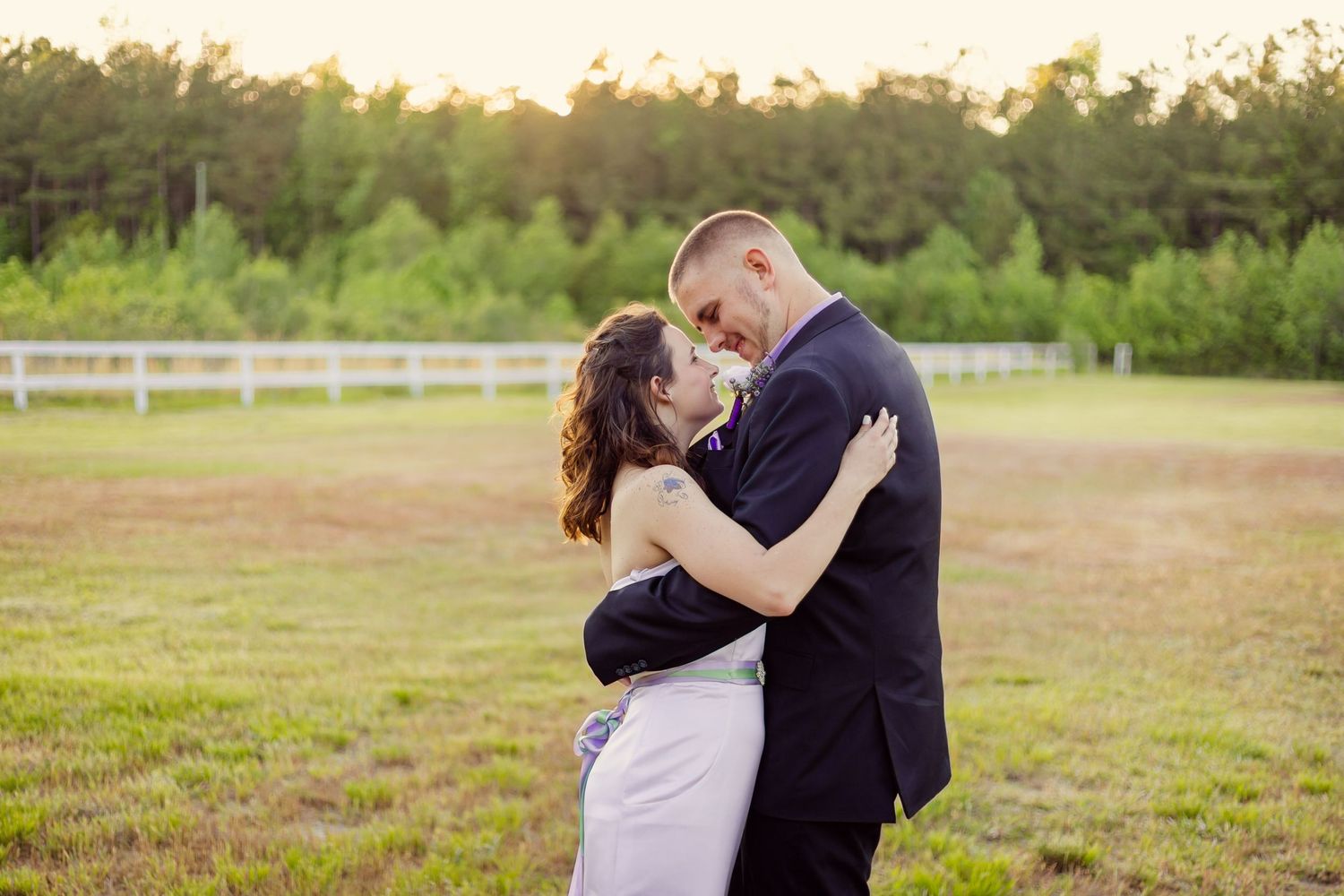 Newlyweds embrace and smile at each other in a sunlit green field during outdoor wedding photos.