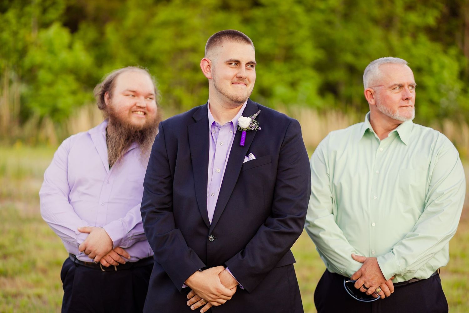 Groom in navy suit with purple boutonniere stands between two groomsmen at outdoor wedding.
