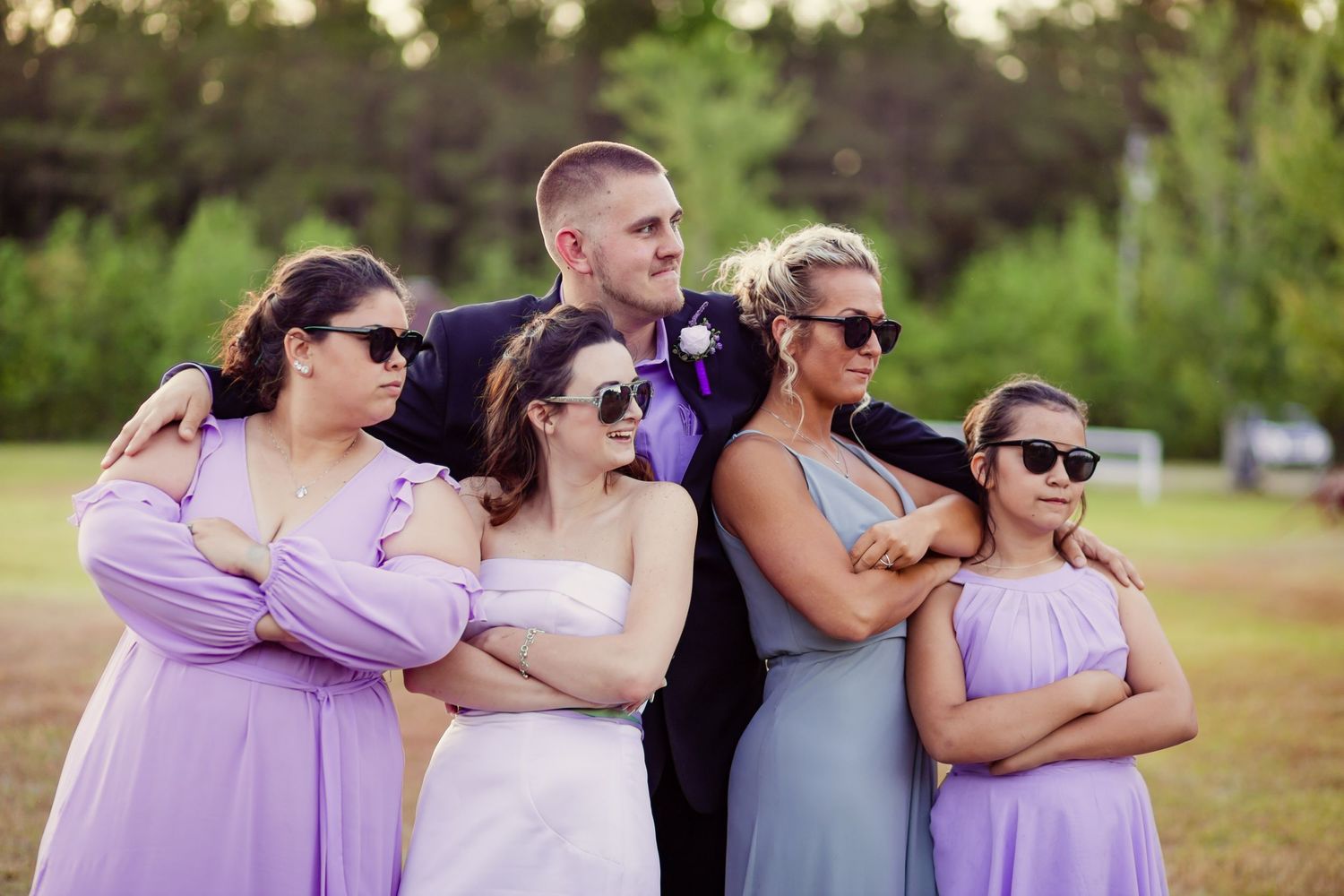 Groom poses with bridesmaids in lavender dresses wearing sunglasses in a fun outdoor wedding photo.