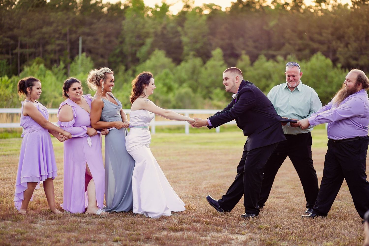 Wedding party plays tug of war on grassy field, bridesmaids in lavender dresses versus groomsmen.