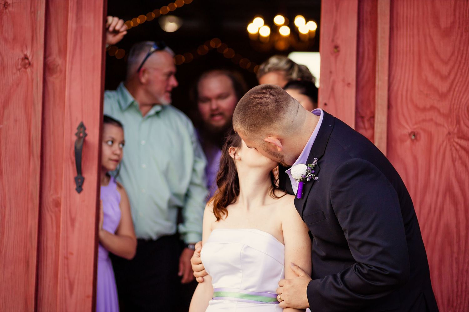 Newlyweds kiss in front of rustic red barn doors while wedding guests watch and celebrate.