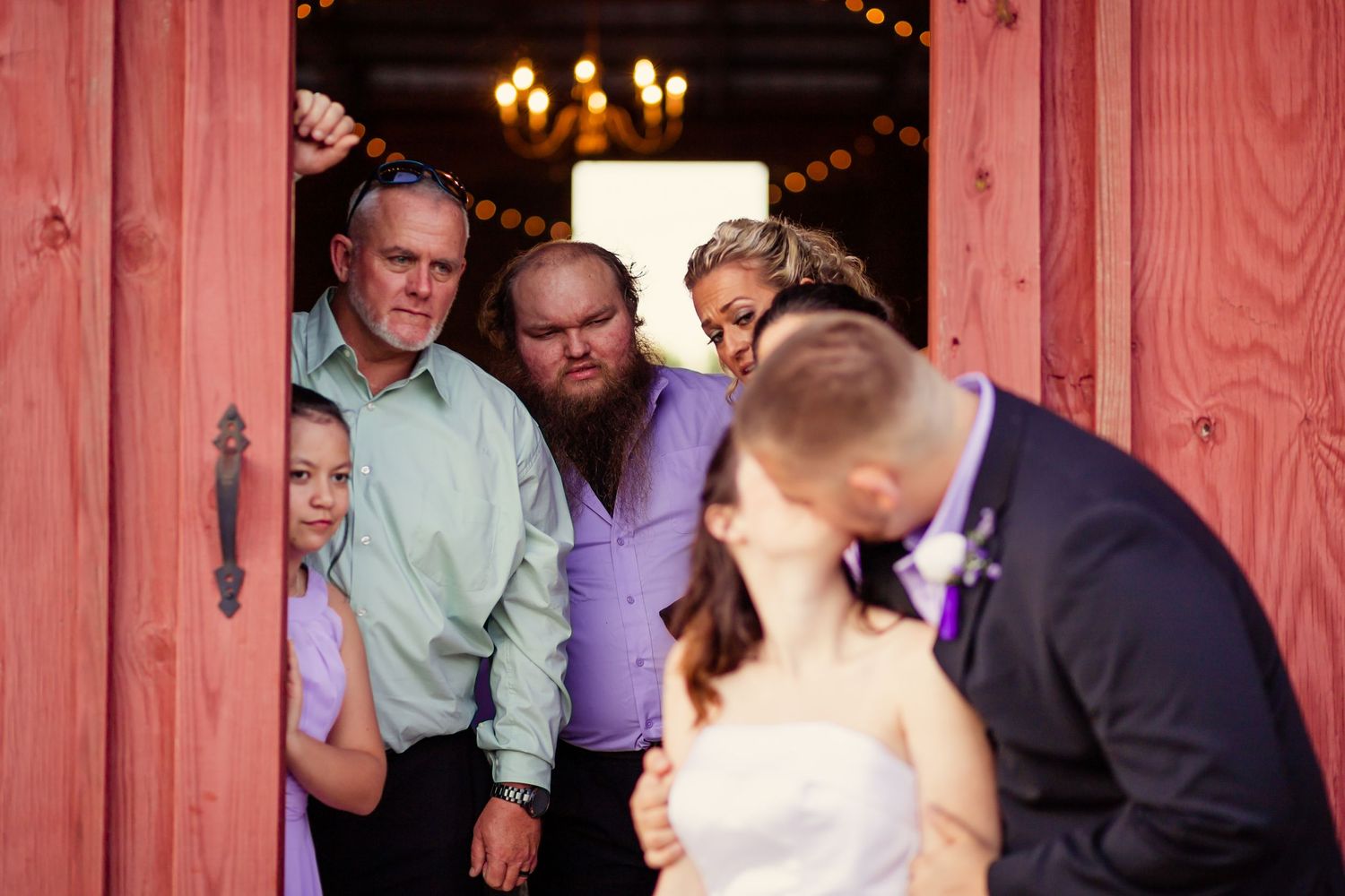 Wedding guests peek through red barn doors watching bride and groom share a kiss outside.