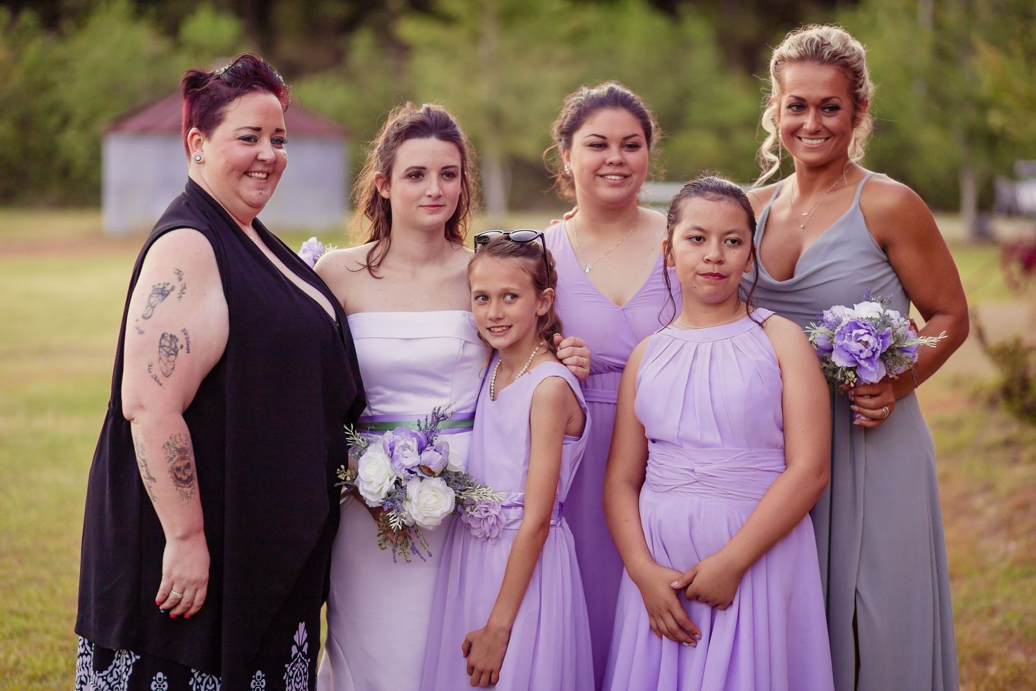 Bridesmaids in lavender dresses pose with bride holding white bouquet at outdoor wedding ceremony.