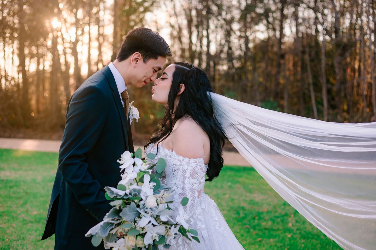 Newlyweds sharing a romantic forehead touch outdoors at golden hour, bride holding a white floral bouquet with veil flowing.