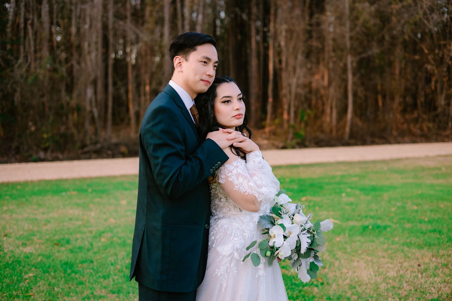 Bride and groom posing intimately in a wooded outdoor setting, bride holding a white eucalyptus bouquet in a lace gown.