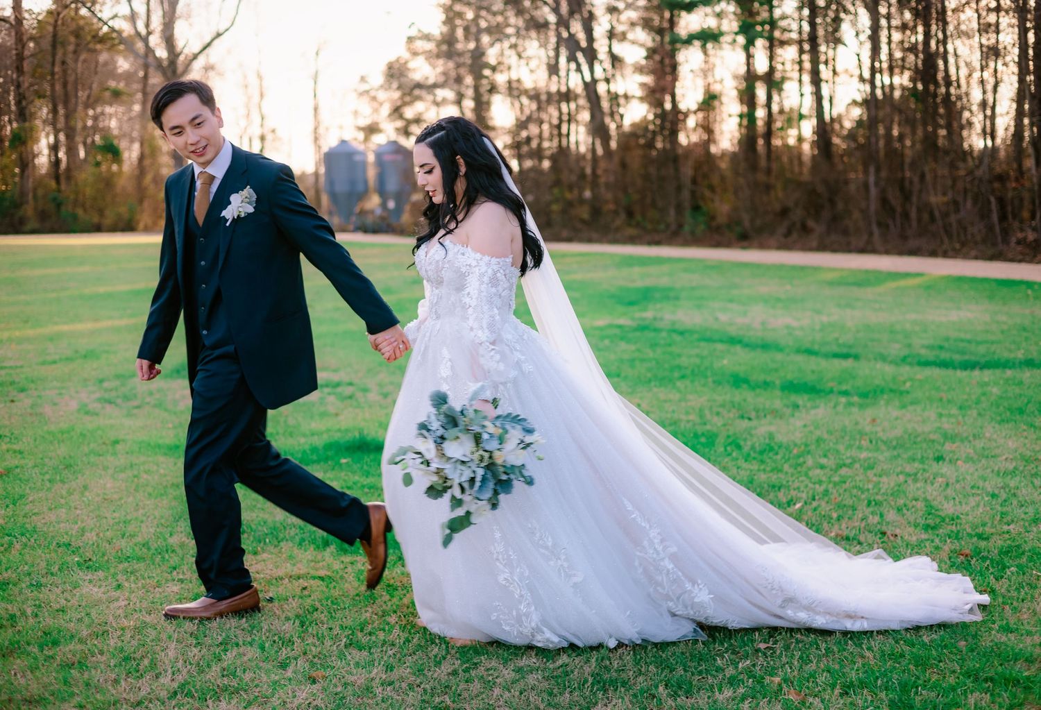 Bride and groom walking hand in hand on green lawn, bride in lace off-shoulder gown with flowing cathedral veil.