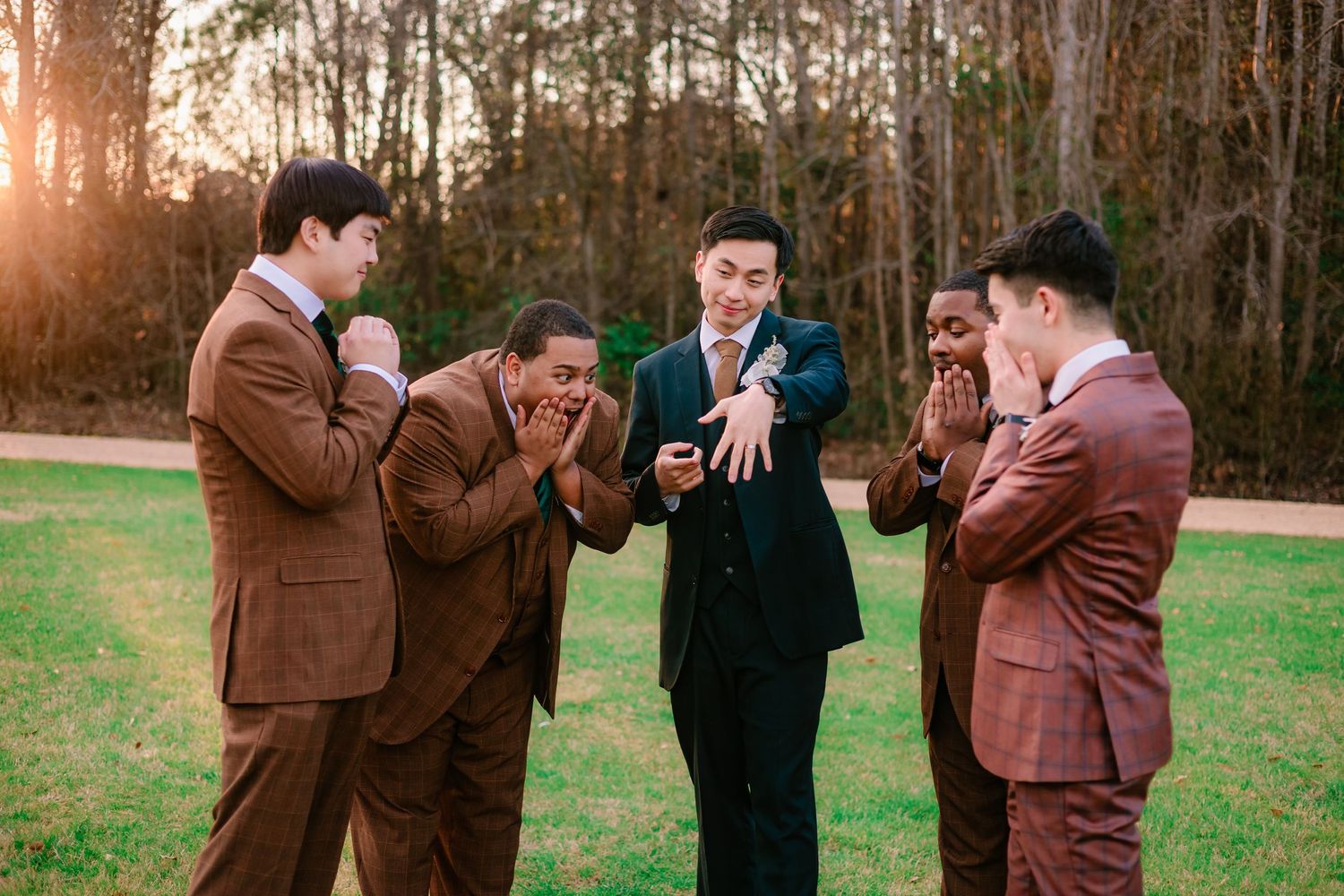 Groom showing his wedding ring to excited groomsmen in brown and burgundy suits during golden hour outdoors.