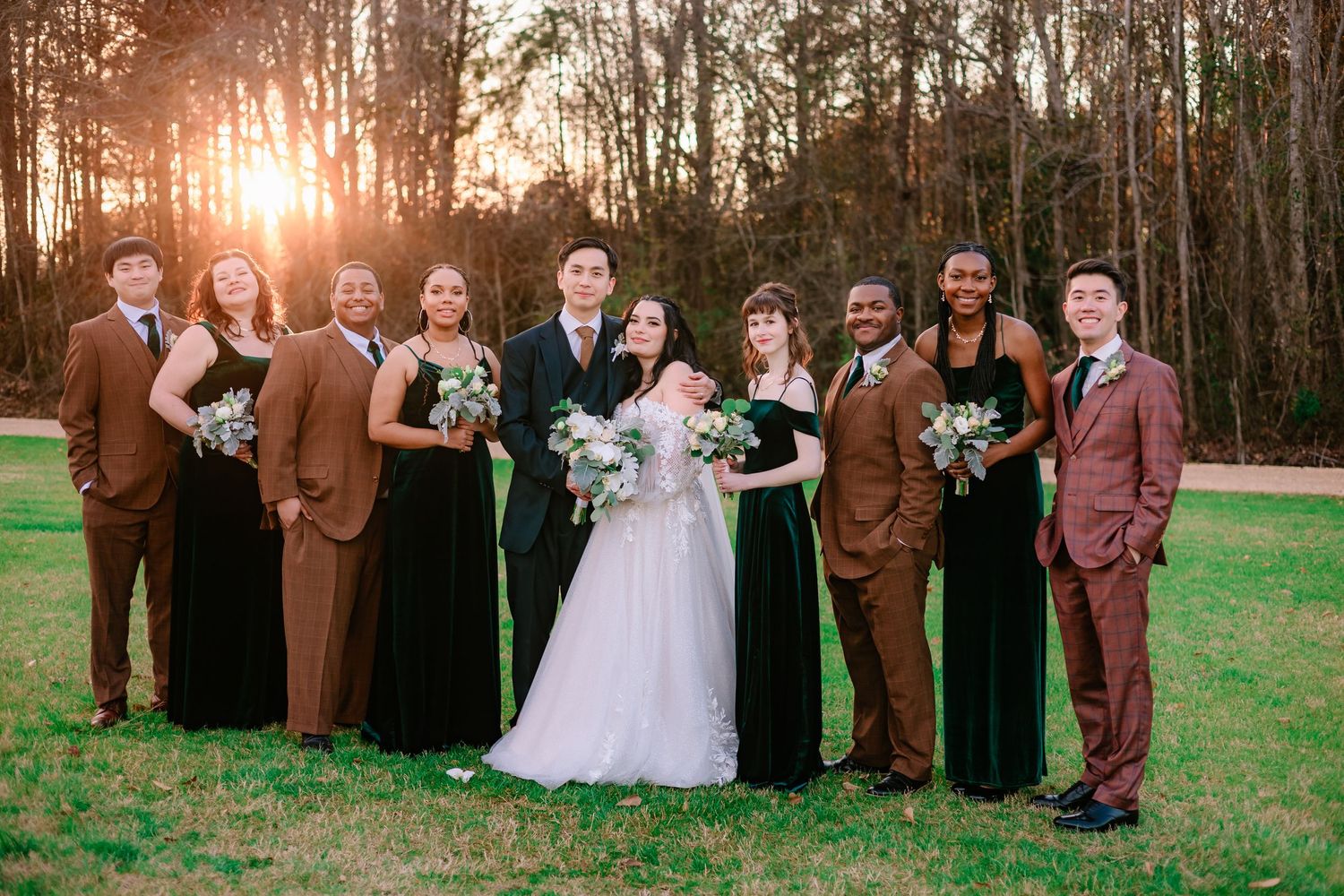 Wedding party posing at sunset, bridesmaids in green and brown velvet dresses with groomsmen flanking the bride and groom.