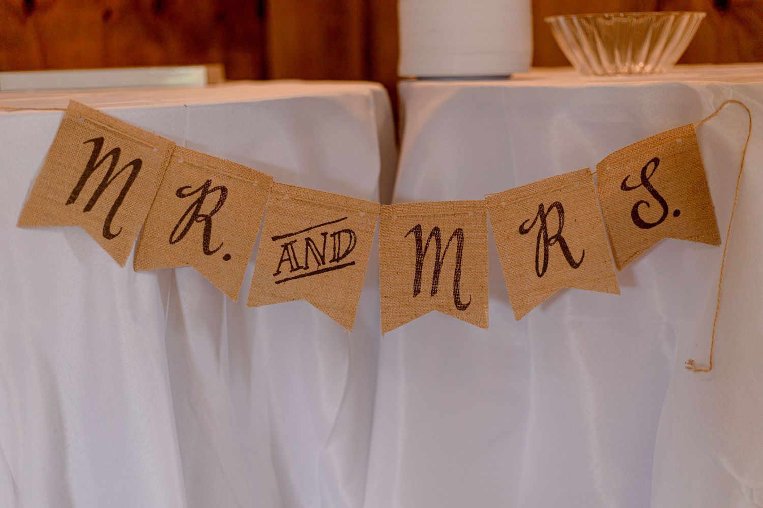 Rustic burlap Mr. and Mrs. banner hanging on white tablecloth at wedding reception.