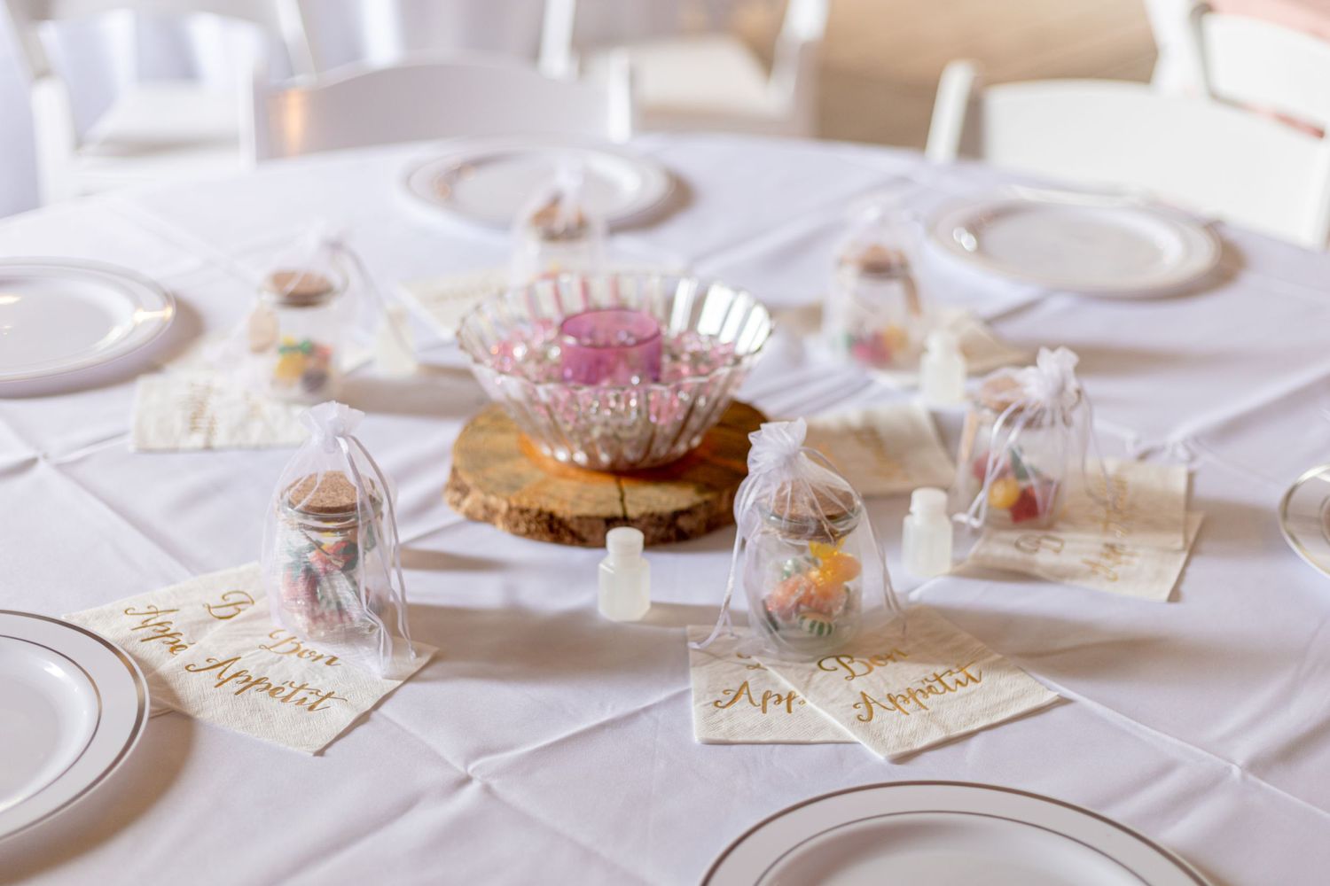 Wedding reception table set with white plates, floral centerpiece, and decorative favor bags.
